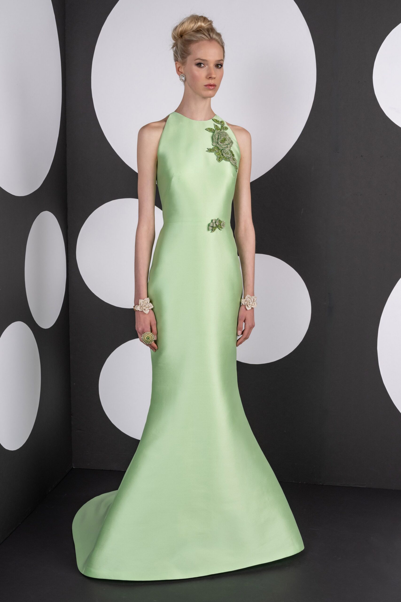 Pale pistachio satin column gown, halter-cut and flaring into a fishtail hem. Beaded floral sprays climbing one shoulder, a smaller bloom pinned at the waist. Matching blossom cuffs at both wrists. Oversized rosette ring with concentric beading. Crystal cluster studs. Garden wedding reception, the cake not yet cut.