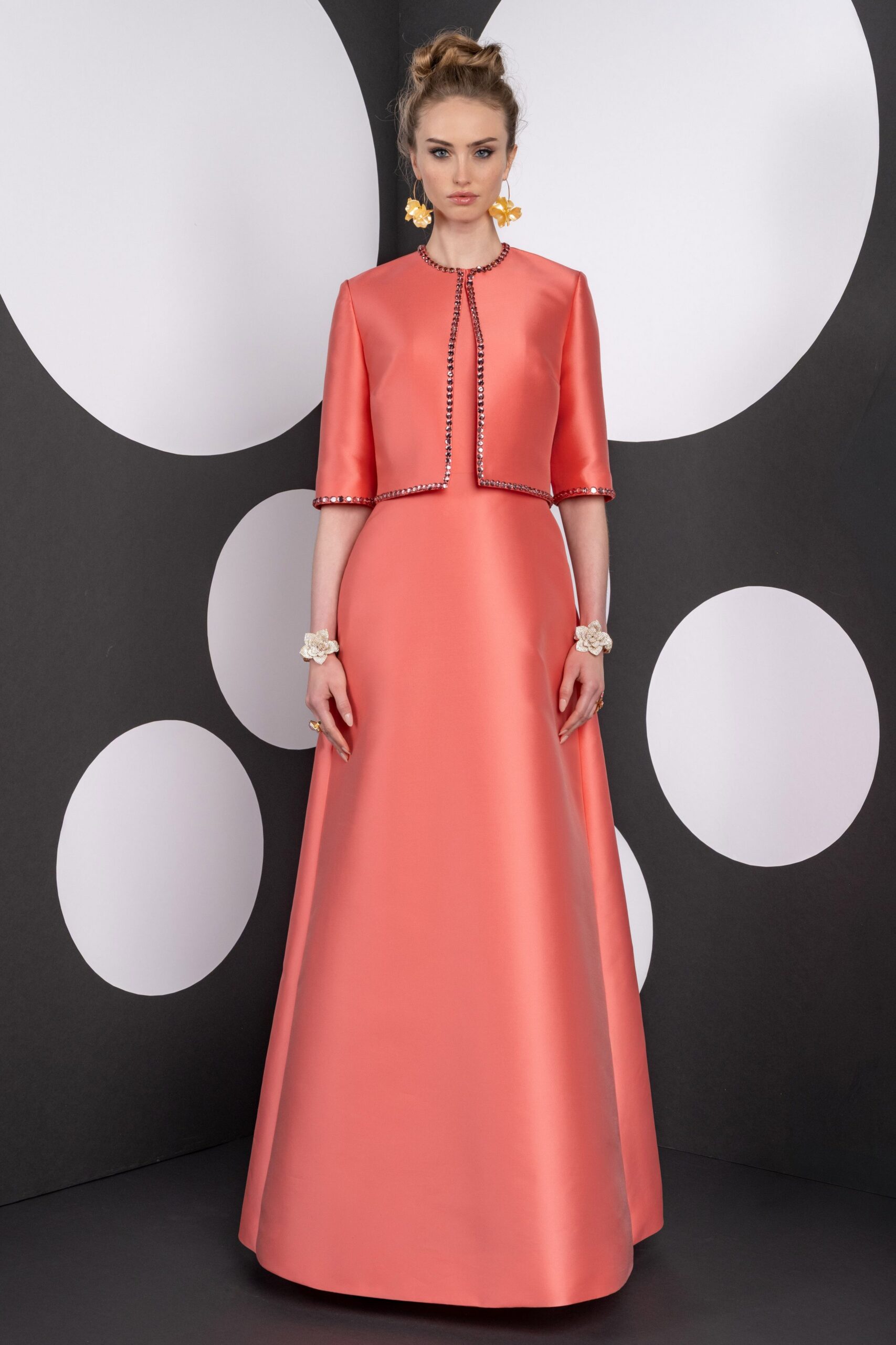 Oversized yellow flower drops brushing the jaw. Coral satin bolero, crystal trim running the round neck and edges. Matching floor-length satin column, widening gently from a high waist seam. Pearly blossom cuffs at both wrists. At a spring luncheon where the tablecloths are brighter than the walls.