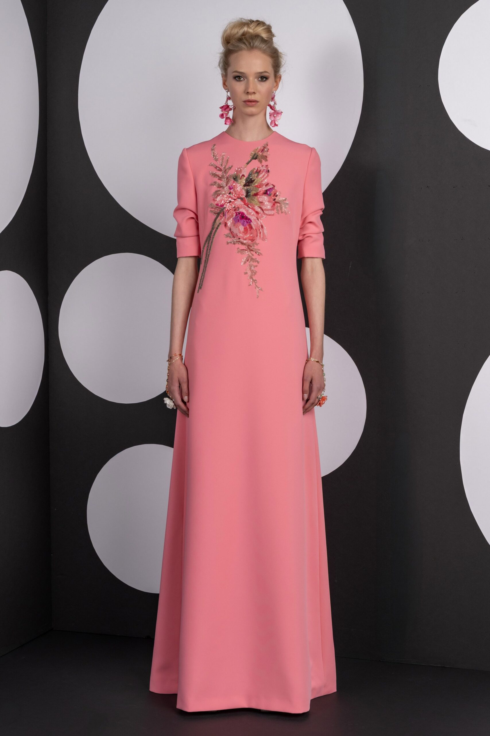Bubblegum pink column gown with bracelet sleeves and beaded flowers trailing off-center. Dangling pink petal clusters. Fine chain bracelets at both wrists. Beaded flower rings, one white and one coral. Garden wedding reception after sunset, champagne sweating onto the linen.