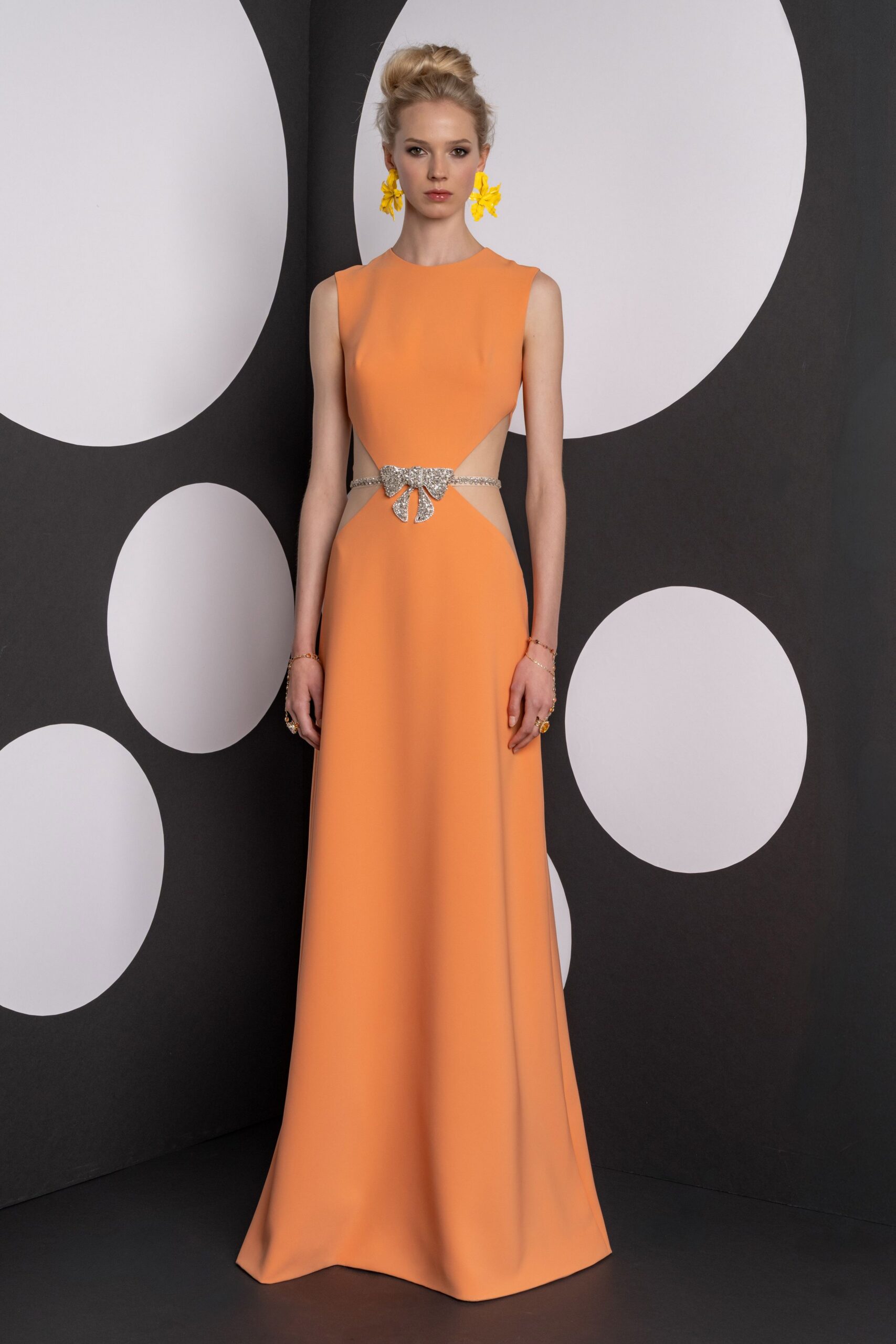 Apricot floor-length gown with deep side cutouts and a crystal bow at the waist. Oversized marigold flower drops. Fine chain bracelets looping to rings at each hand. Garden party after sunset, standing near the last tray of sliced melon.