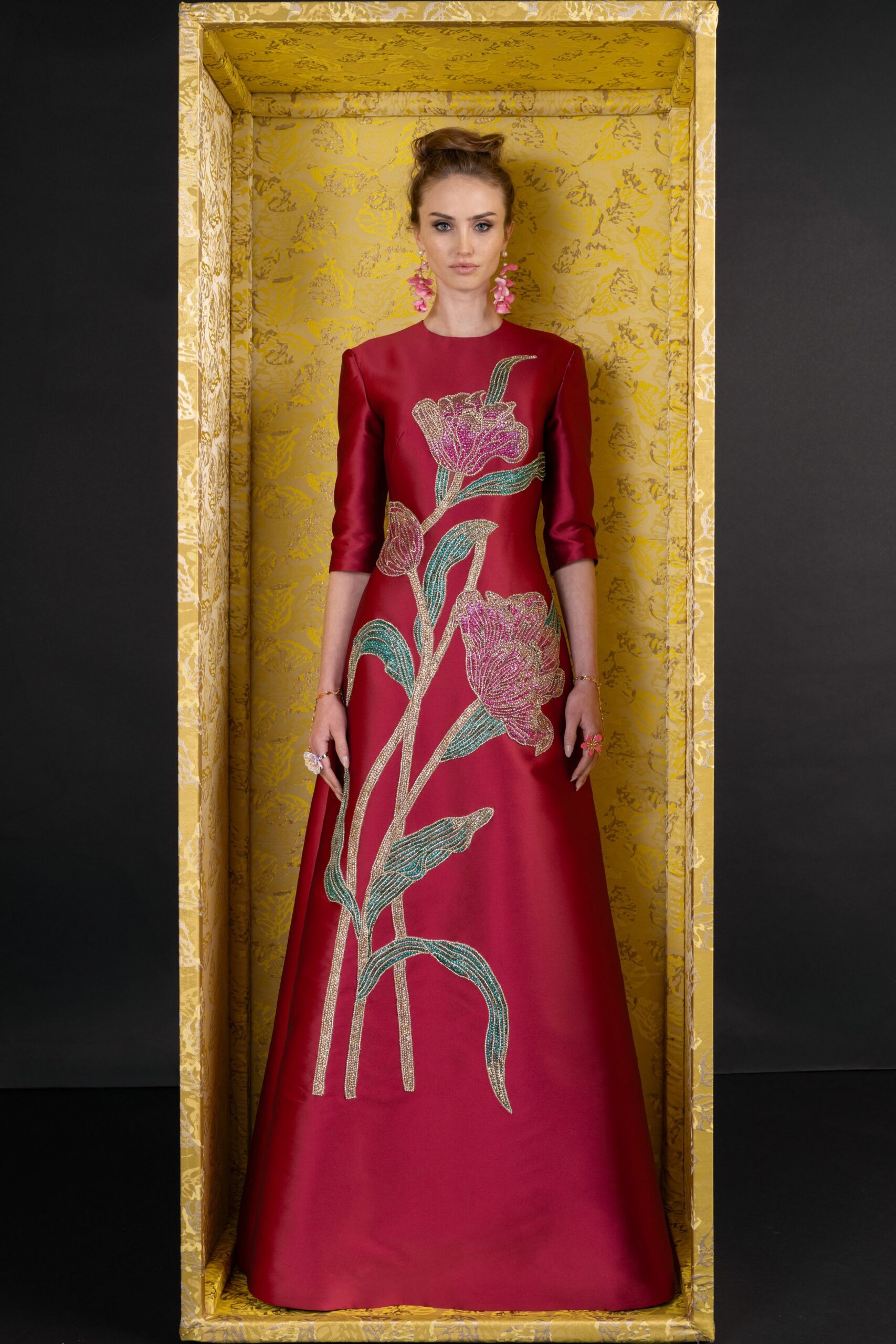 Crimson satin column gown with elbow sleeves and giant beaded tulip stems. Pink blossom cluster drops. Fine gold chain bracelets. Oversized floral cocktail rings. At the museum fundraiser, standing inside the shipping crate before the auction starts.