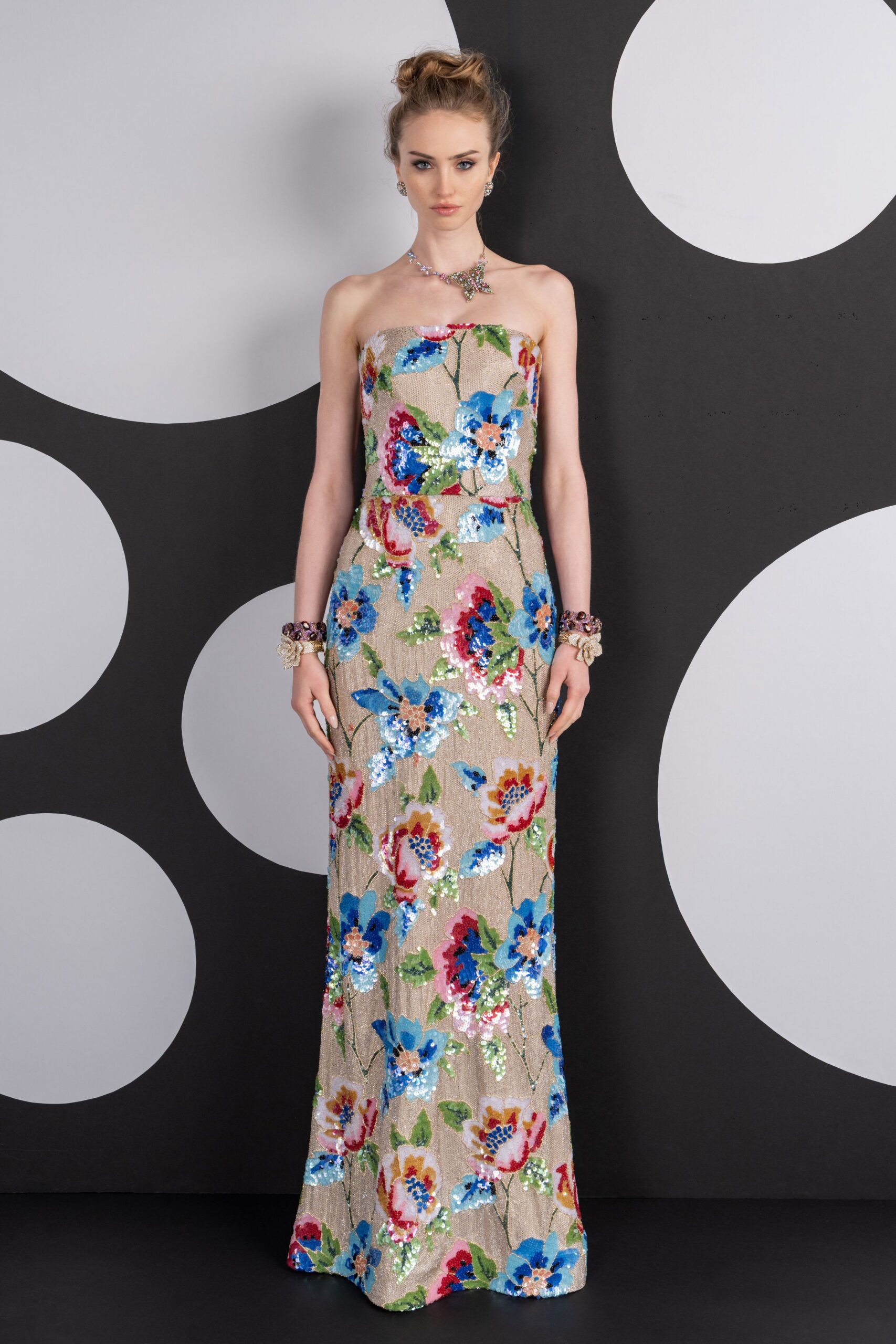 Asymmetric crystal floral necklace skimming one collarbone. Round clustered crystal studs. Strapless column gown in nude mesh, drenched with blue and pink sequined flowers. Chunky gem cuffs over pale lace wrist corsages. Garden wedding reception after dark, standing near the cake before the first cut.