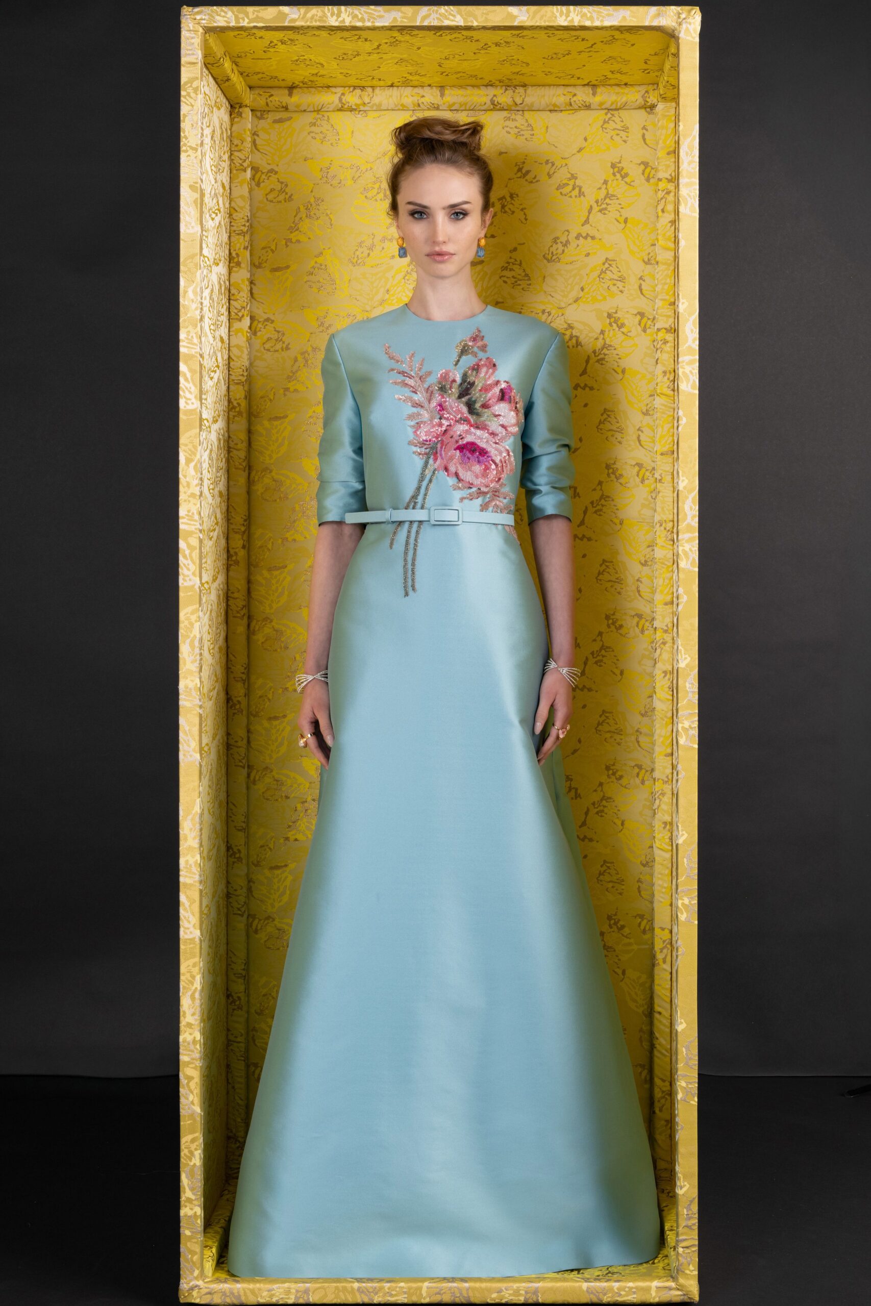 Pale aqua satin gown with elbow sleeves and sequined peony embroidery. Narrow self belt with a covered rectangular buckle at the waist. Blue drop earrings with gold bead tops. Silver wire bracelets stacked at both wrists. In a wallpapered drawing room before the toast turns into a speech.