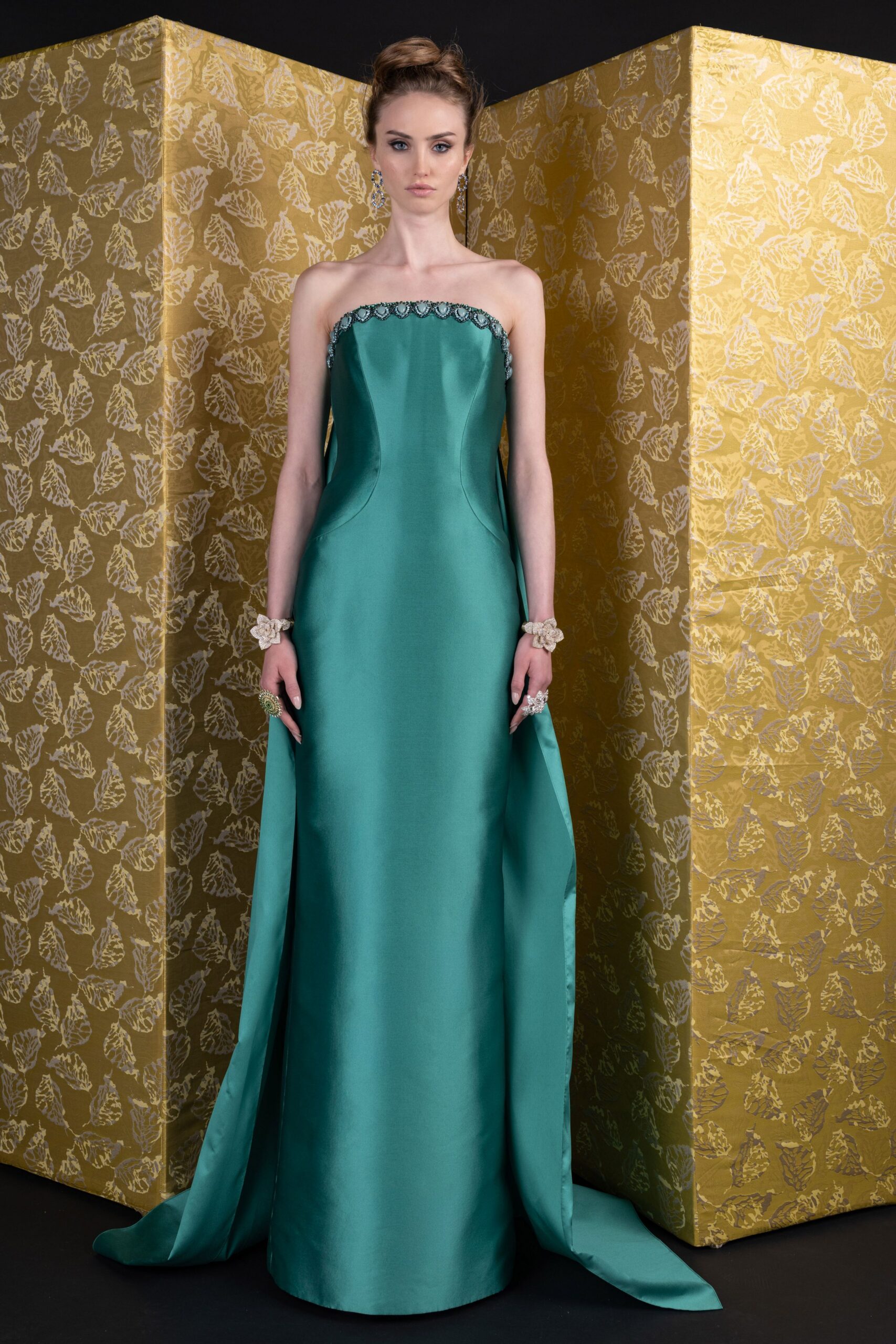 Dangling crystal cluster earrings brushing the jaw. Strapless sea-green satin column gown, scalloped beading tracing the straight neckline. Chunky floral bracelets at both wrists. Oversized crystal rings catching light at each hand. At the top of a hotel staircase before the string quartet starts.