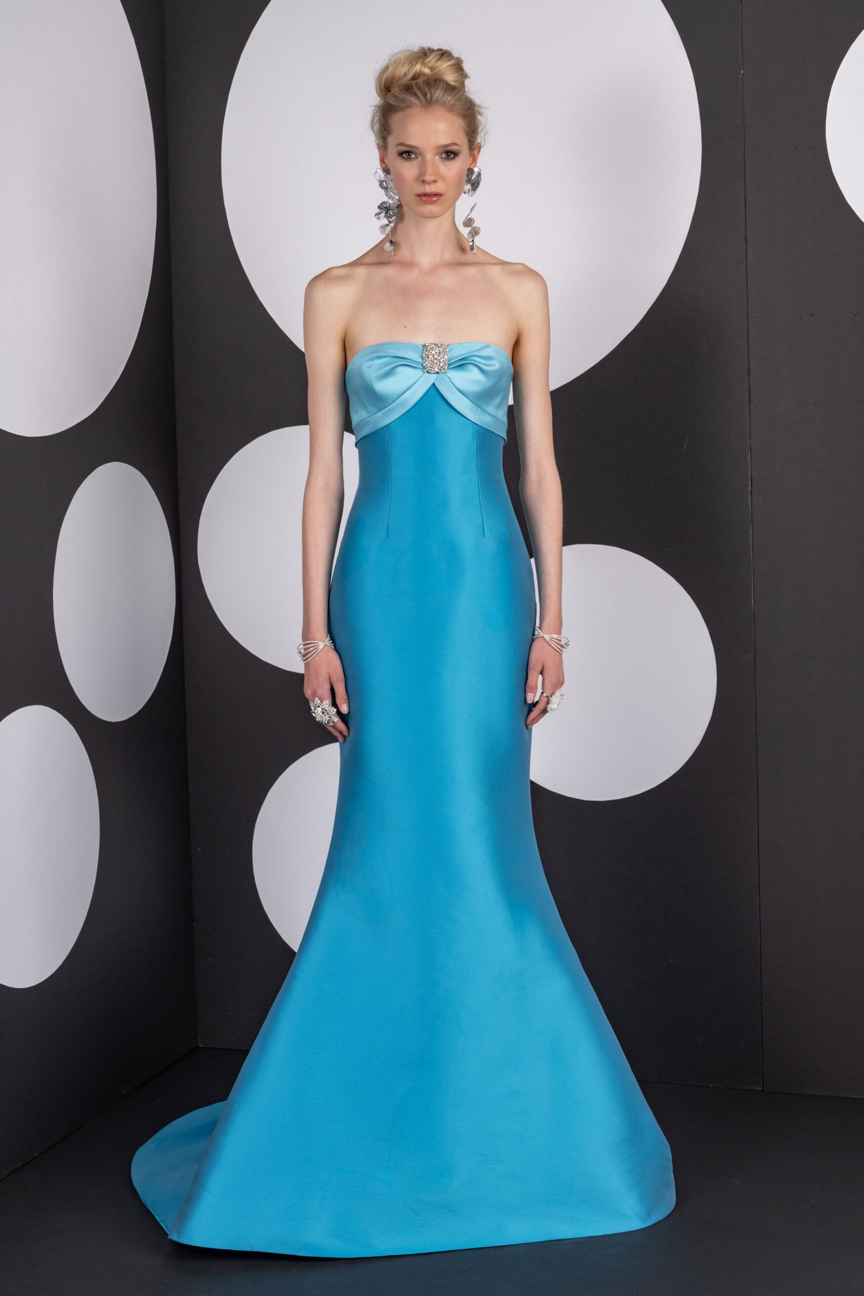 Long crystal flower drops brushing the collarbones. Strapless aqua satin column, ruched bust pinned with a square rhinestone clasp. Silver coil cuffs stacked at both wrists. Clustered crystal rings catching at two fingers. Hotel ballroom carpet before the charity auction starts.