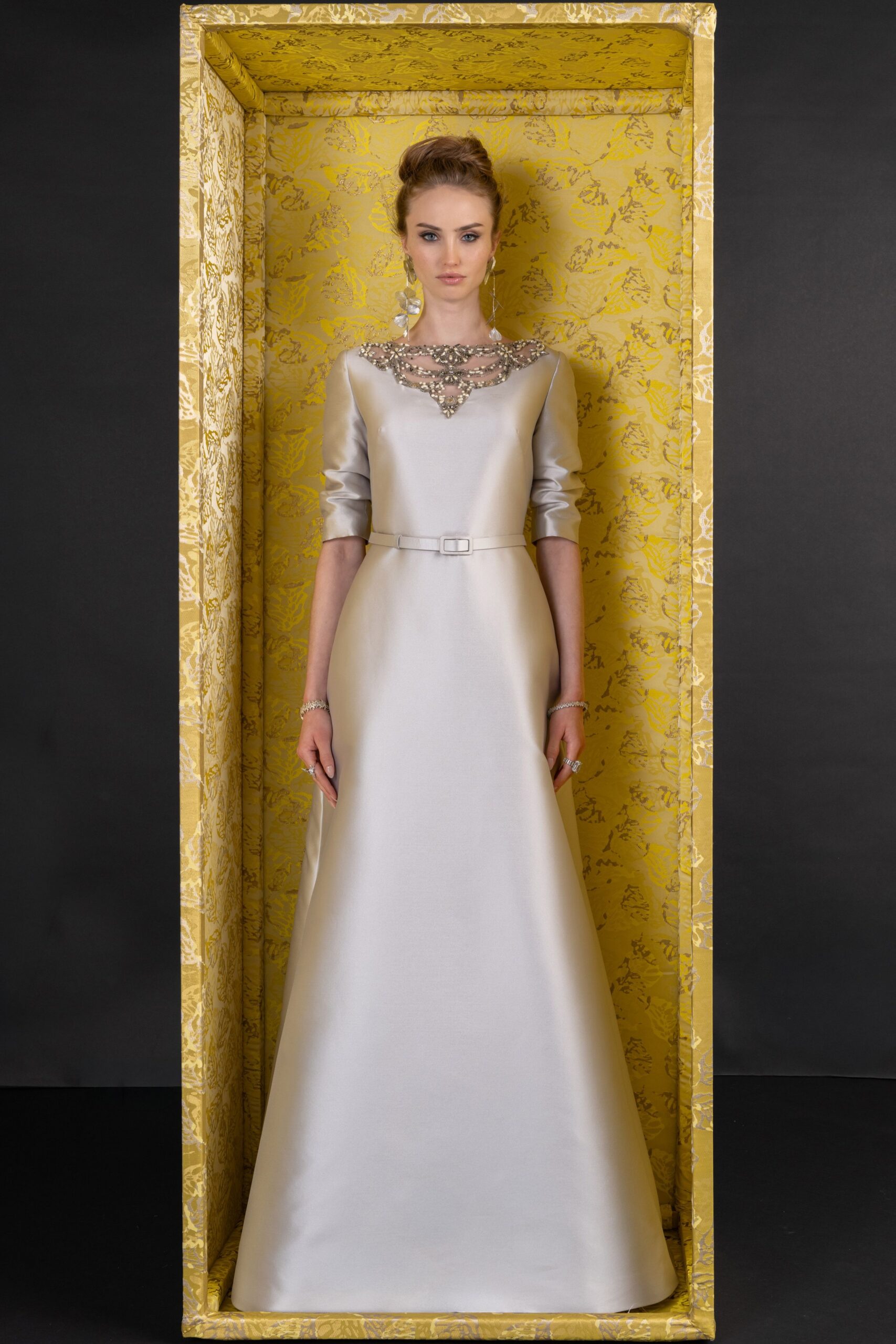 Ivory duchess satin gown with a crystal lattice collar and three-quarter sleeves. Narrow self-belt with a small rectangular buckle at the waist. Long drop earrings with translucent petal-shaped stones. For greeting relatives in a hotel suite before the wedding cars arrive.