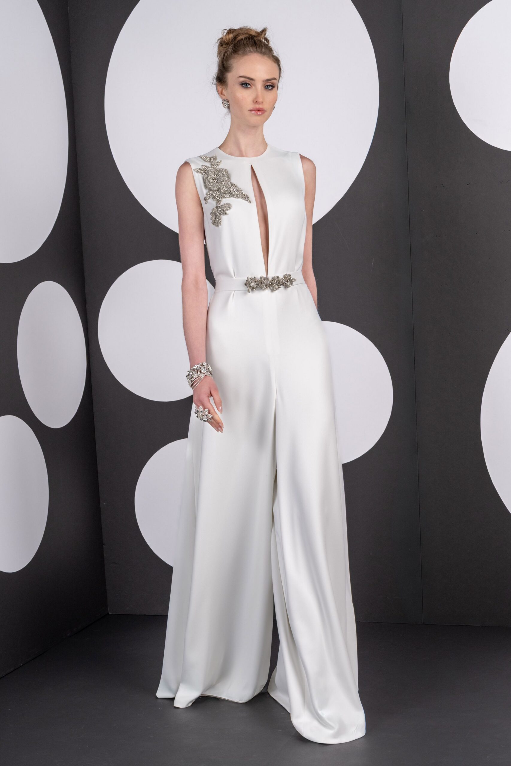 Ivory crepe jumpsuit with a plunging slit and crystal floral appliqué. Clustered silver beading cinching the waist seam. Round crystal cluster studs. Stacked silver cuffs with twisted strands. Oversized crystal flower ring. Registry office stairwell, bouquet paper still crackling under one arm.