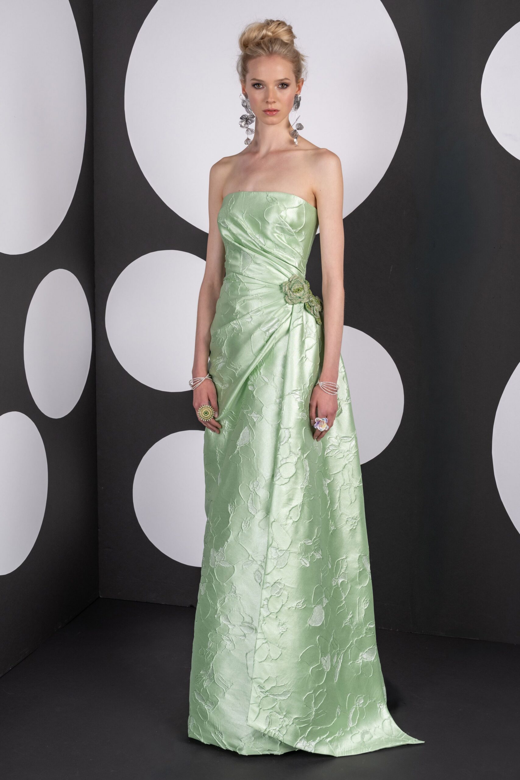 Long silver petal clusters swinging nearly to the shoulders. Strapless mint jacquard gown, ruched sideways into a beaded flower at the hip. Coiled silver bangles stacked at both wrists. Oversized flower rings, one pistachio and one lilac. Garden wedding reception, the cake table already losing petals in the heat.