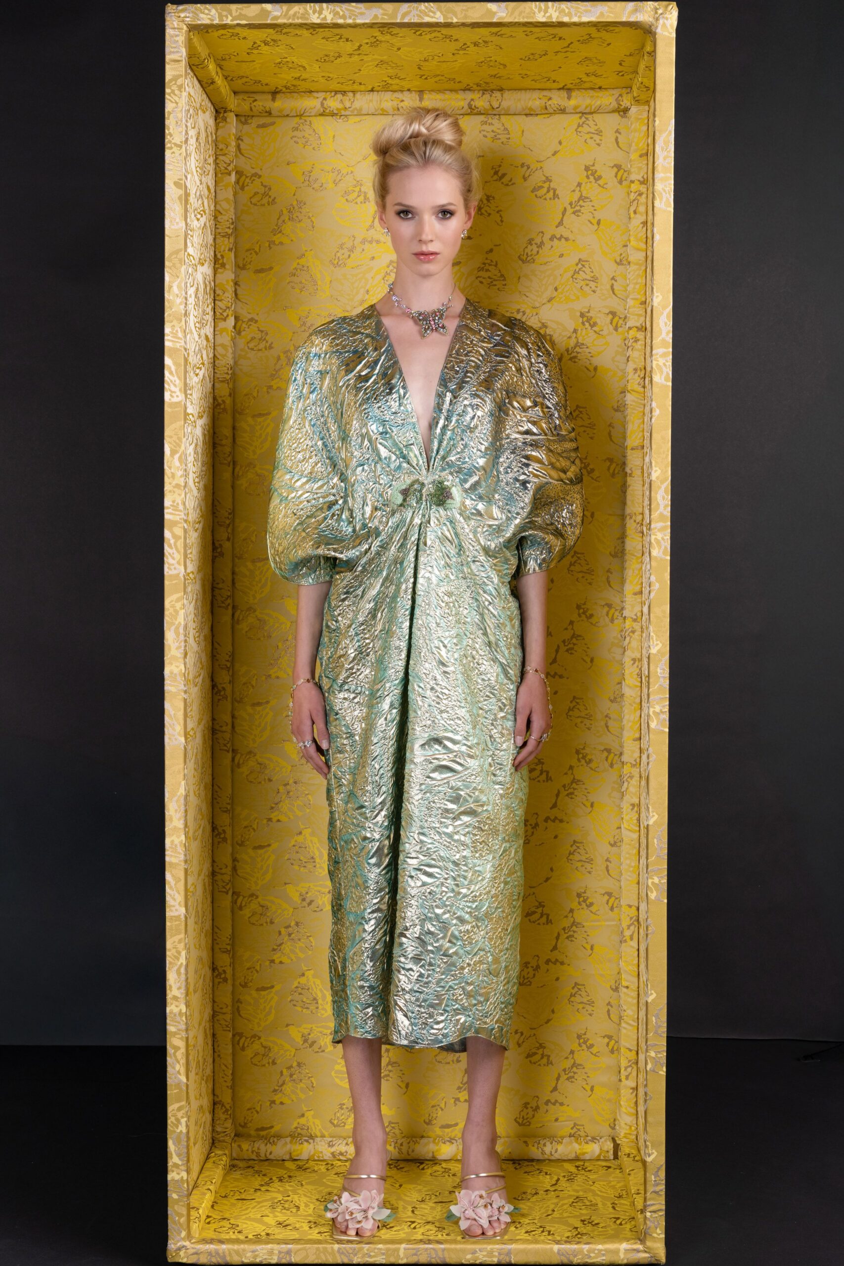 Pastel crystal butterfly necklace sitting high at the collarbone. Seafoam lamé dress, deeply plunged and ruched into a puckered knot. Barely there metallic sandals topped with pale organza flower clusters. Standing inside a wallpapered gift box before the auction starts.