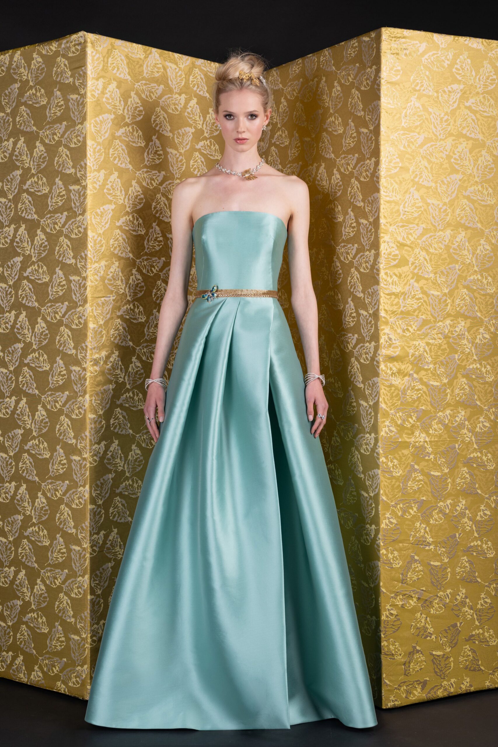 Ice-blue satin strapless ballgown, box pleats opening from a narrow metallic waist. Gold braided belt with a tiny blue jeweled flourish off-center. Coiled crystal bracelets stacked at both wrists. Close-set crystal necklace with a small gold leaf drop. Charity auction in a paneled ballroom, after the speeches and before dessert.