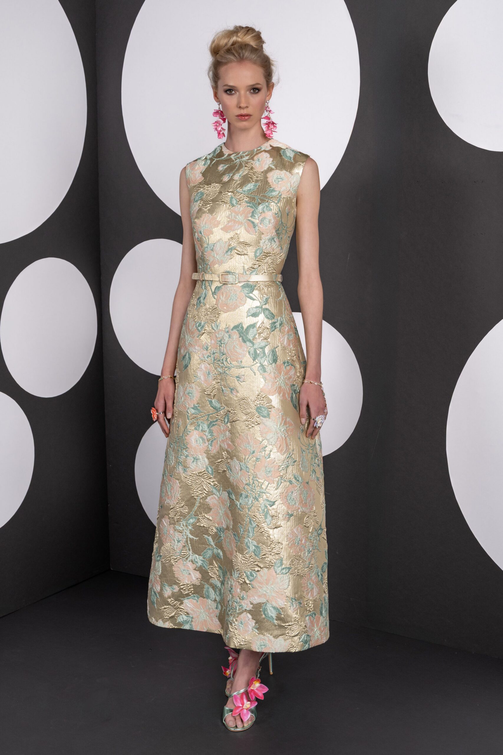 Sleeveless gold brocade dress with blush peonies and pale aqua leaves. Narrow self belt with a small square buckle at the waist. Long pink blossom clusters brushing the jaw. Metallic heeled sandals wrapped with oversized orchid blossoms. Garden wedding reception, cake already cut and shoes sinking into grass.
