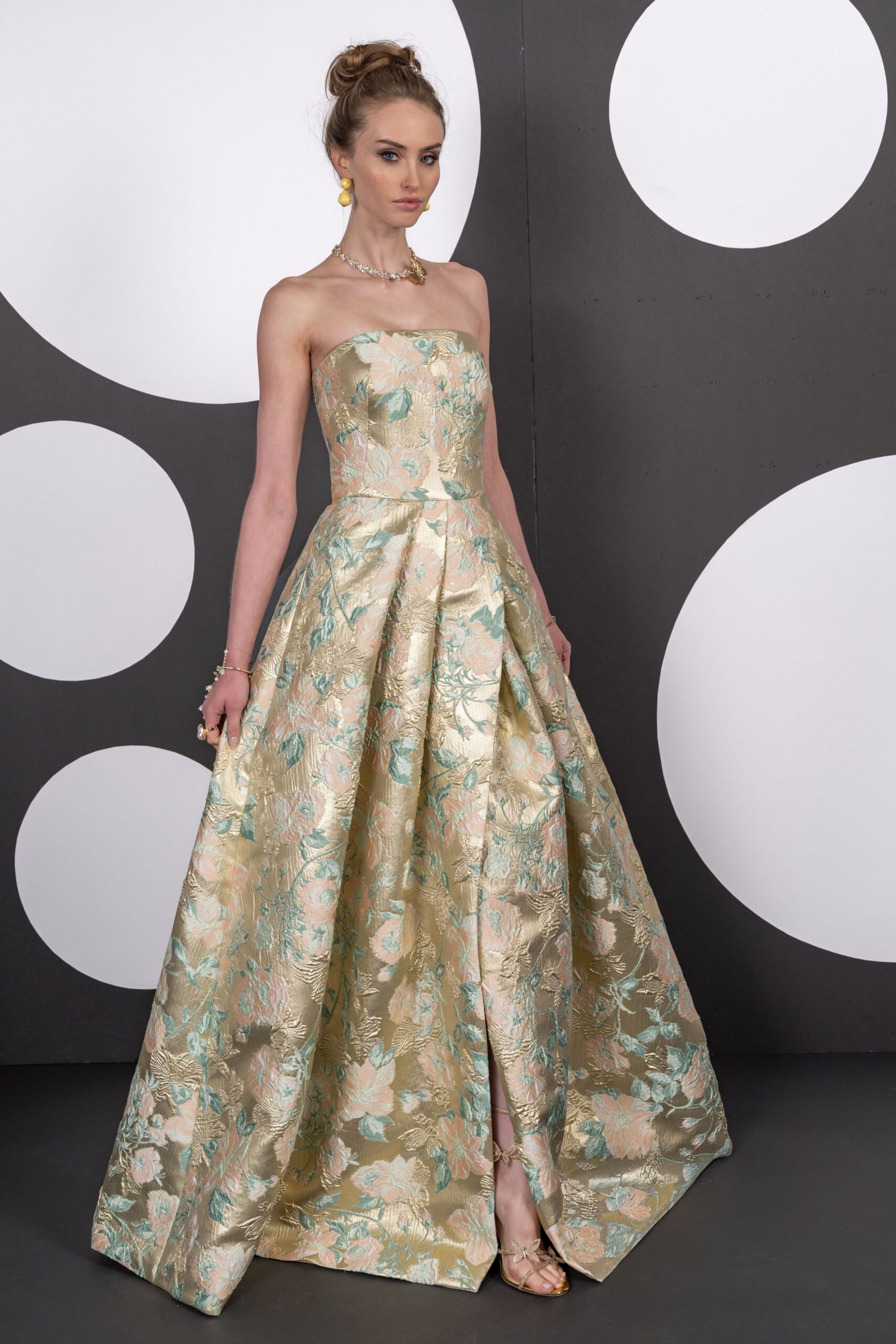 Strapless ball gown in metallic floral brocade, box pleats opening from a dropped seam. Lemon-yellow drop earrings. Crystal strand with one clustered gold flourish. Gold sandals with a tiny bow across the toes. Garden wedding reception, the cake already cut and the grass turning damp.