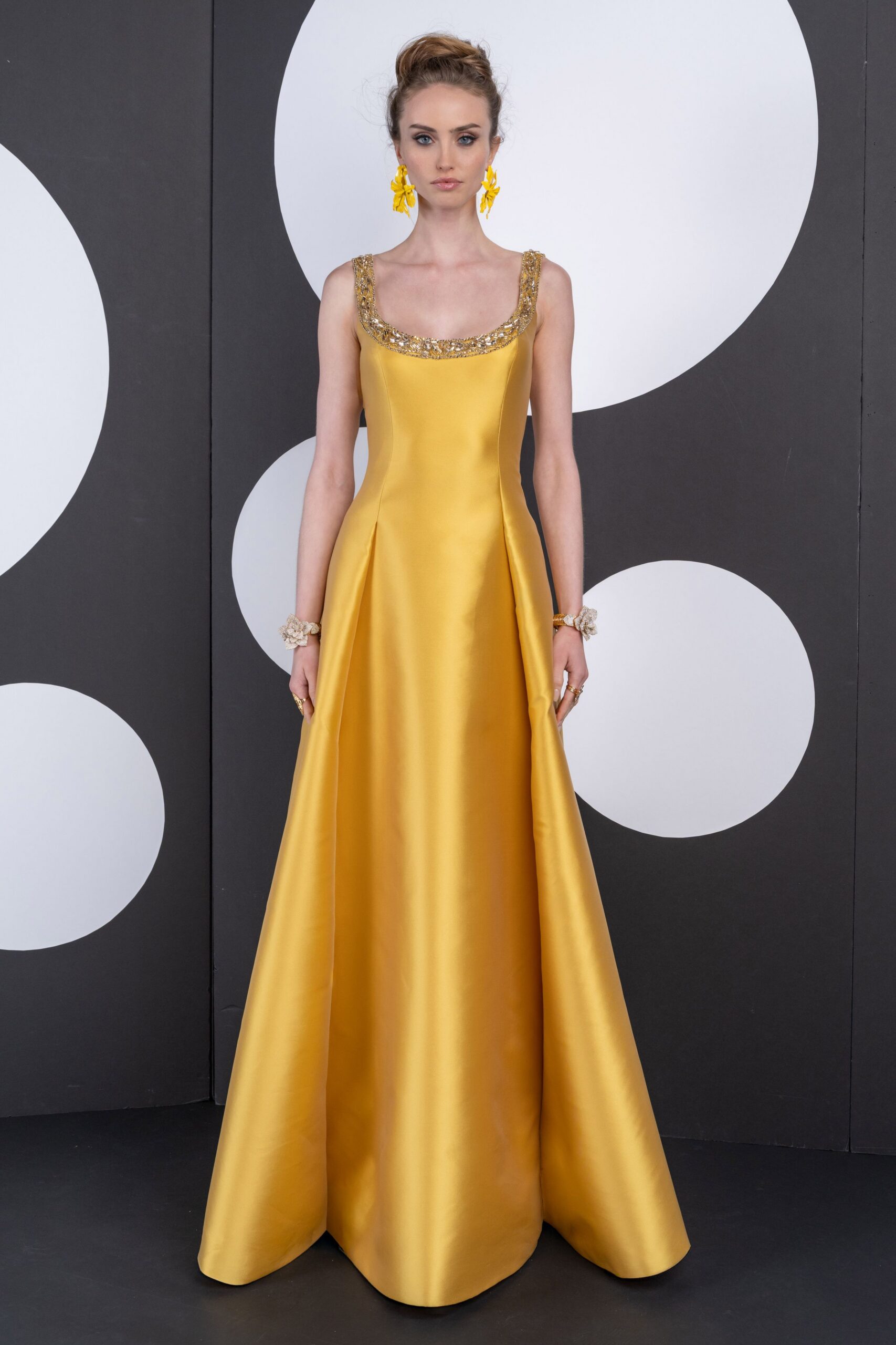 Lemon-yellow flower-drop earrings with layered petal shapes. Marigold satin ball gown, scoop neckline crusted with gold beading. Clustered crystal-flower bracelets at both wrists. Slim gold rings stacked across several fingers. For arriving at a garden wedding just before the light turns honey.