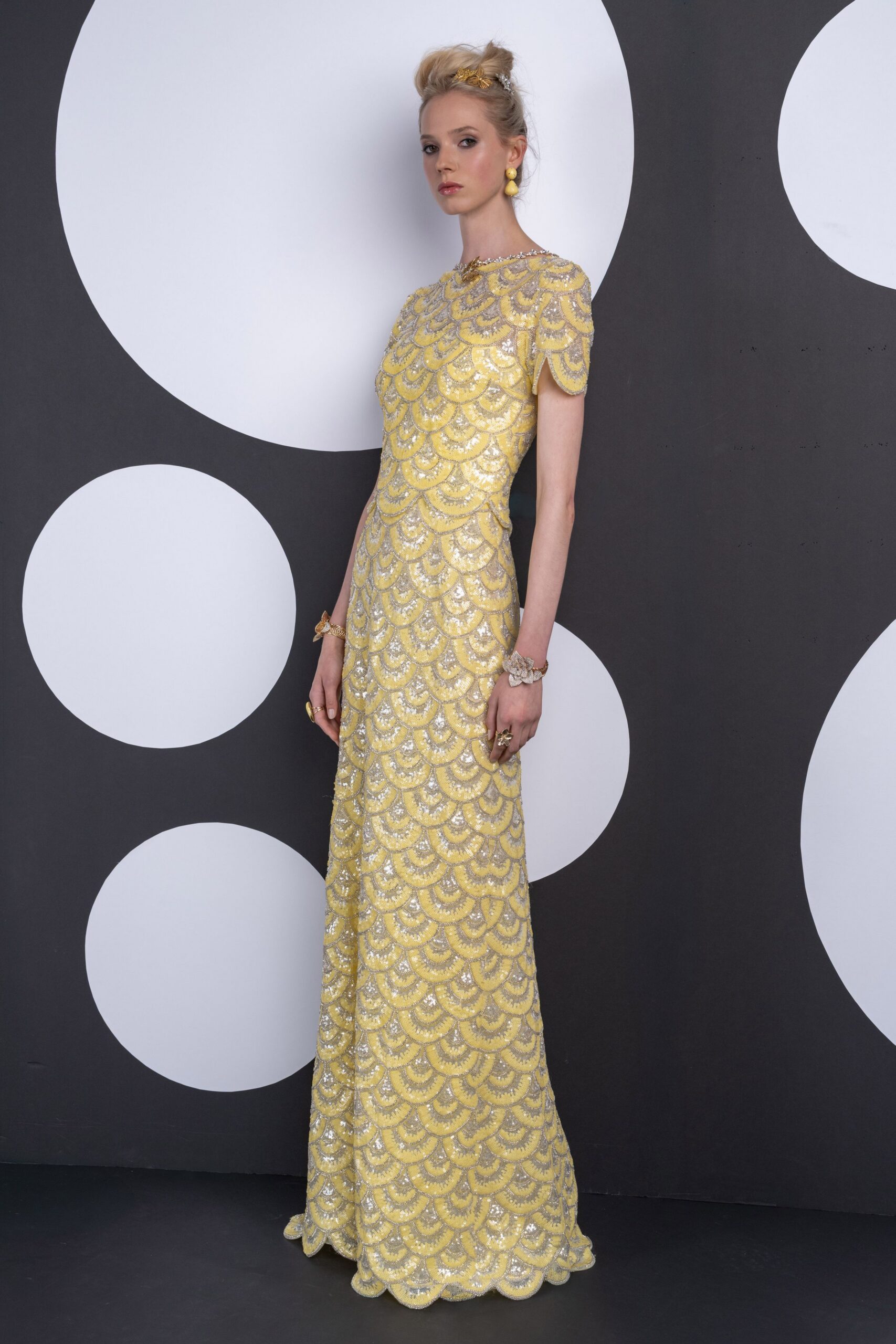 Gold leaf clip pinned into a high, airy twist. Double-drop yellow bead earrings. Floor-length gown in lemon and silver scalloped sequins, cap sleeves and a petal hem. Mismatched floral cuffs at both wrists. Garden wedding reception, champagne already warm on the tray.