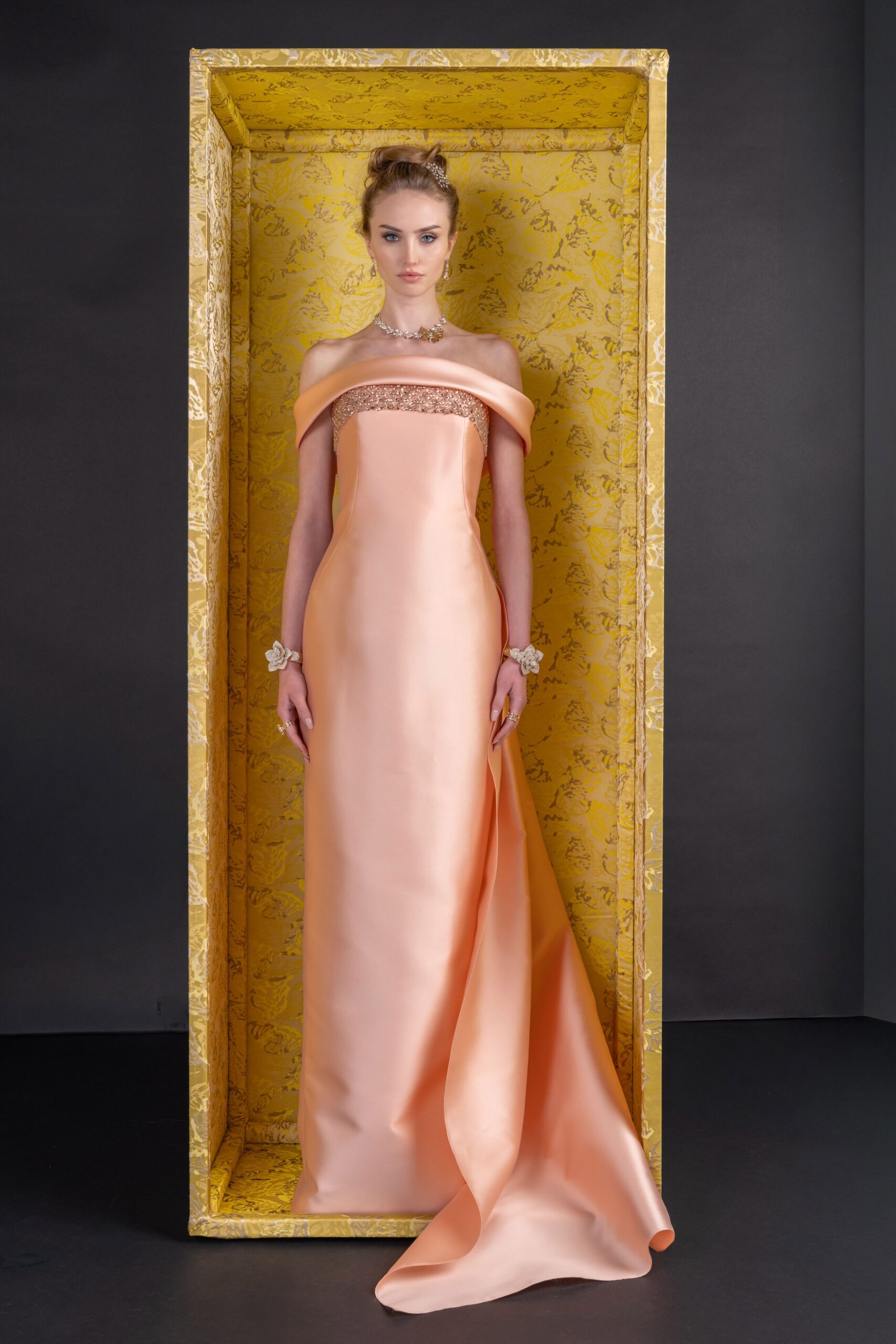 Peach satin column gown with a rolled off-shoulder band and glittering bandeau edge. Crystal cluster choker with one insect-shaped motif at the throat. Matching crystal flower cuffs, one on each wrist. Small clustered rings on several fingers. Standing inside the museum crate before the auction preview opens.