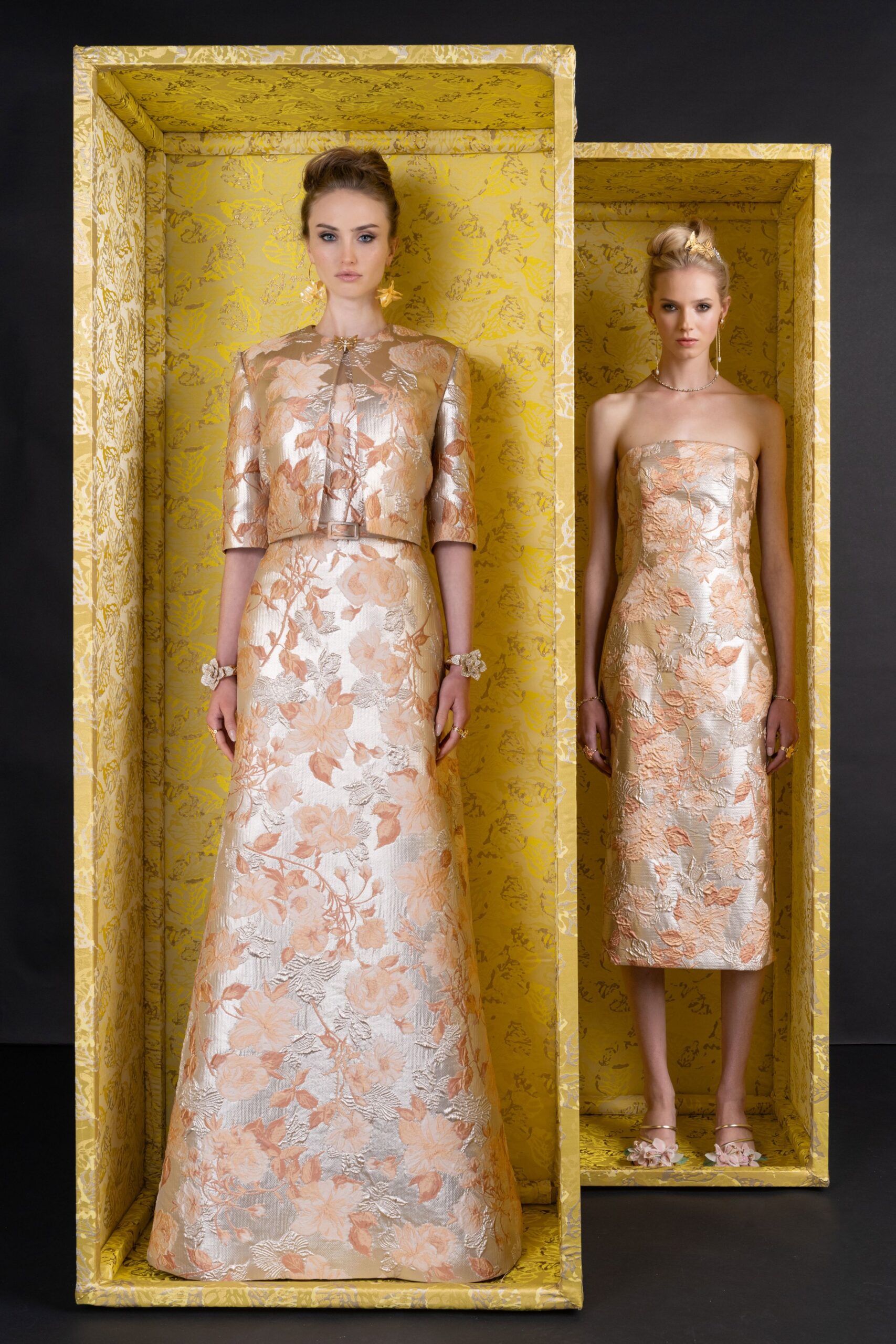 Champagne brocade jacket with apricot florals and a jewel clasp. Matching floor-length A-line skirt, heavy woven sheen holding its shape. Flower corsages at both wrists. Gold starburst drops. In the receiving line before a hotel ballroom wedding finally starts.