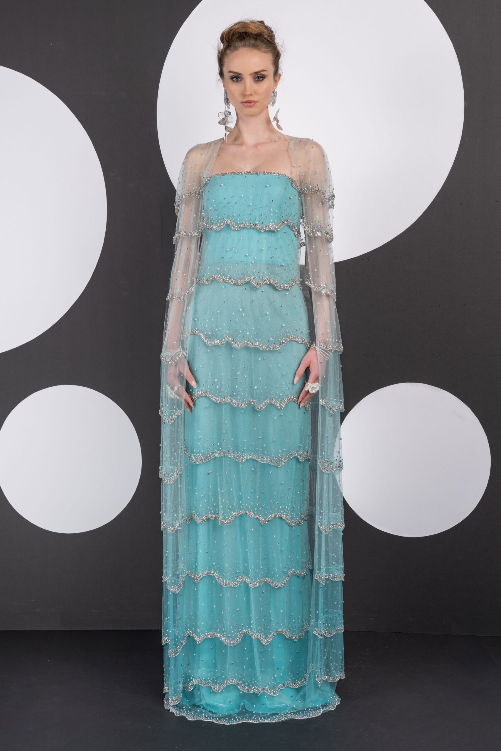 Seafoam tulle column gown with scalloped crystal bands tiered to the floor. Sheer cape sleeves hanging from the shoulders, beaded at every wavering edge. Long translucent flower drops. Cluster flower ring at one hand. Hotel ballroom after midnight, collecting champagne bubbles in the hem.