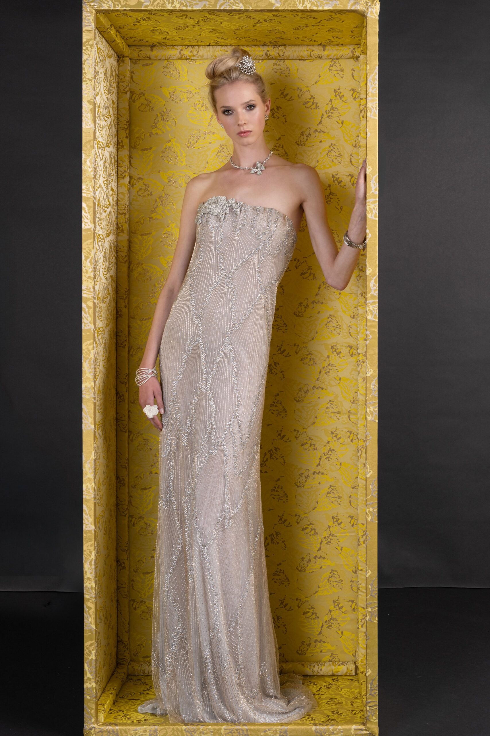 Crystal spray pin tucked into a smooth high twist. Rhinestone necklace with a small cross-shaped drop at center. Strapless blush column gown densely beaded in fanburst and vine lines. Stacked crystal bangles on both wrists. Oversized white flower ring. At the hotel florist before the ballroom doors open.