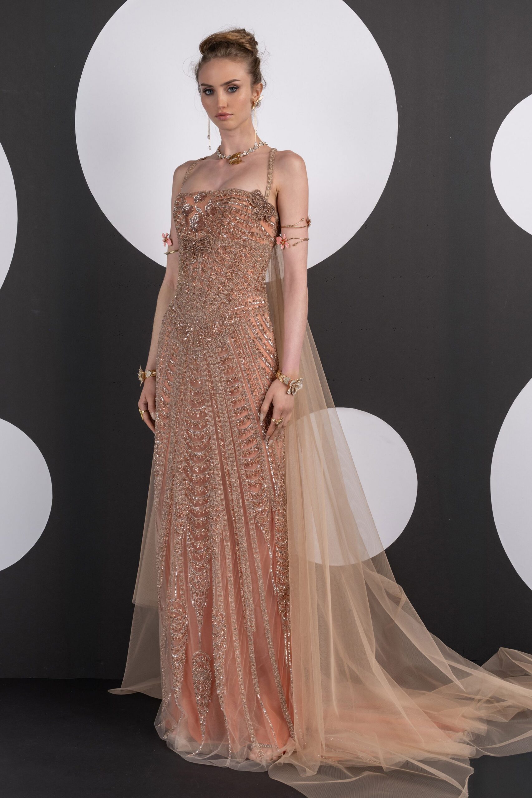 Blush column gown crusted in silver beading, with bow motifs at the bust. Nude tulle train falling from the straps and pooling far behind. Gold spiral arm cuffs tipped with tiny pink flowers. Barely there metallic sandals under the hem. For the hotel ballroom hour when the string lights finally come on.