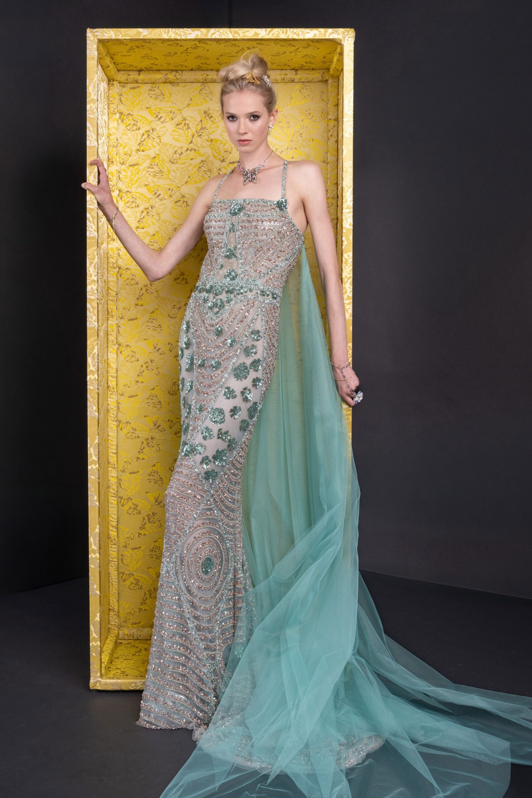 Nude column gown densely beaded in mint scrolls and concentric arcs. Seafoam tulle panel falling from the back into a long gauzy sweep. Crystal butterfly at the throat. Fine chain bracelets climbing the wrist. Large flower ring catching the fingertips. At a winter charity auction beneath hotel chandeliers and too much perfume.
