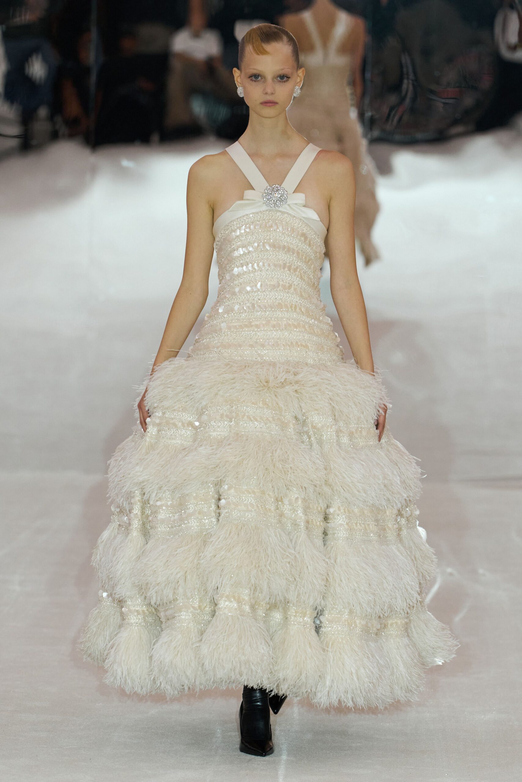 Ivory ballgown banded with sequins and plush feathered tiers, crystal brooch at the crossed straps. Round crystal cluster earrings. Black pointed boots peeking from the hem. At the hotel ballroom wedding where the cake arrives on a mirrored cart.