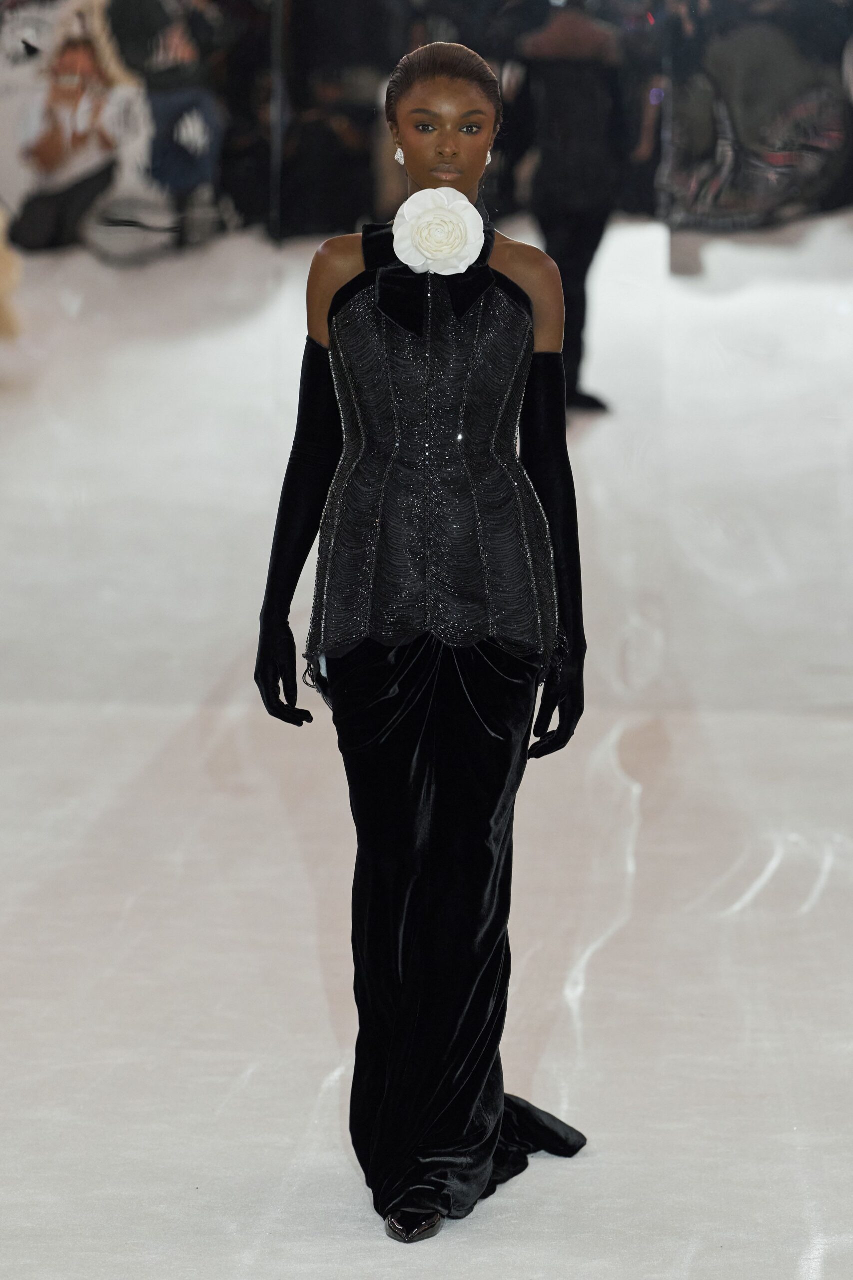 Oversized white camellia pinned tight at the throat. Black velvet column gown with a glittering beaded corset overlay. Opera-length black velvet gloves disappearing under the bare shoulders. Teardrop crystal studs. Black pointed pumps peeking from the hem. Walking across a hotel ballroom before the auction starts.