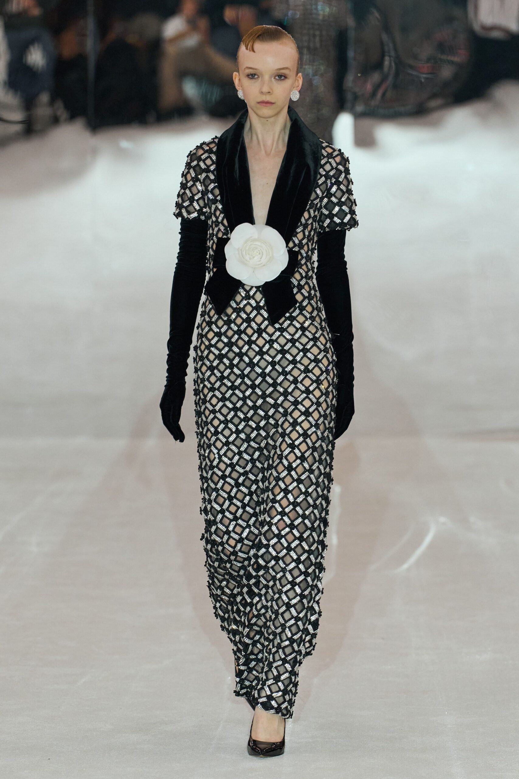 Round crystal cluster earrings. Black lattice-sequin jumpsuit over nude lining, plunging velvet lapels and a white camellia at the waist. Opera-length black velvet gloves, lightly ruched at the forearm. Black patent pumps with a narrow square toe. Hotel ballroom carpet muffled under fresh snow and too much champagne.