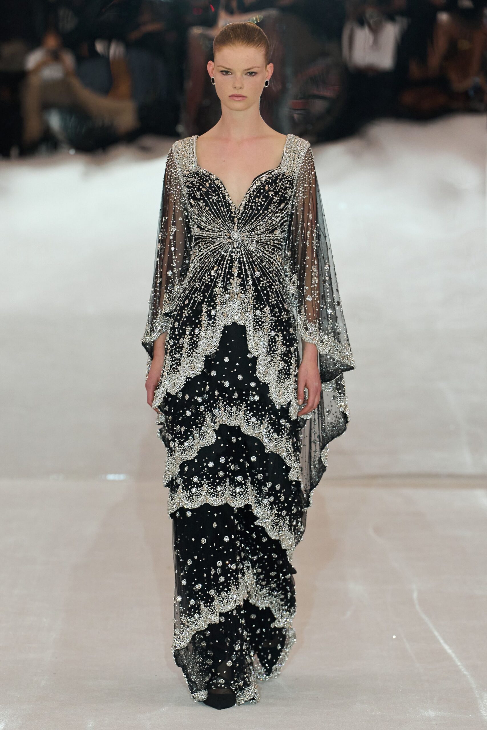 Small black hoop earrings. Black sheer-mesh gown drenched in silver beading, starbursting from the sternum. Cape sleeves falling to handkerchief points, cuffs crusted white with crystals. Hotel ballroom after midnight, the last ice sculpture still sweating.