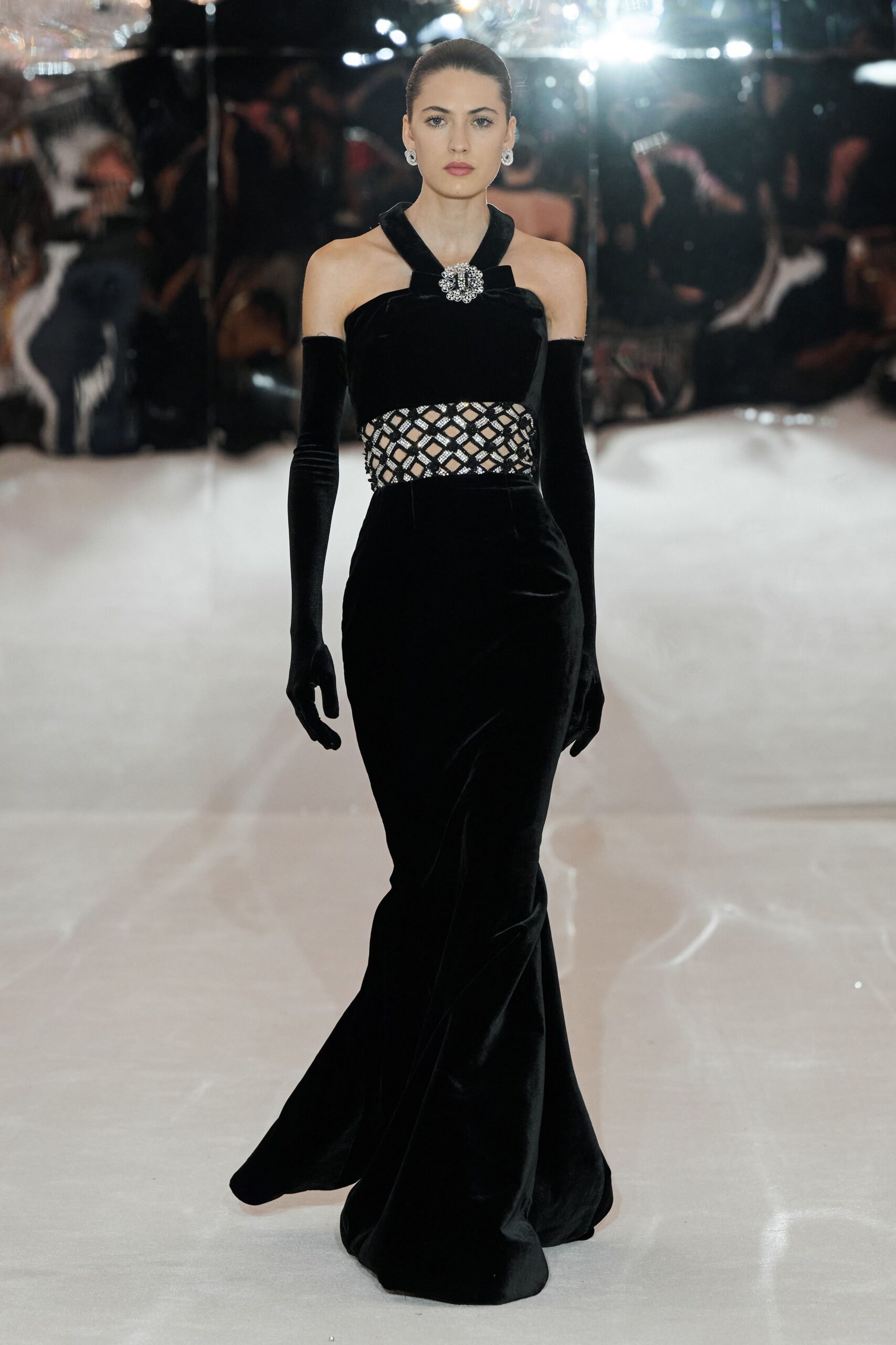 Round crystal cluster earrings. Black velvet halter gown, rhinestone buckle at the throat. Diamond openwork band crusted with silver beading. Black velvet opera gloves. Crossing a hotel ballroom before the auction starts.