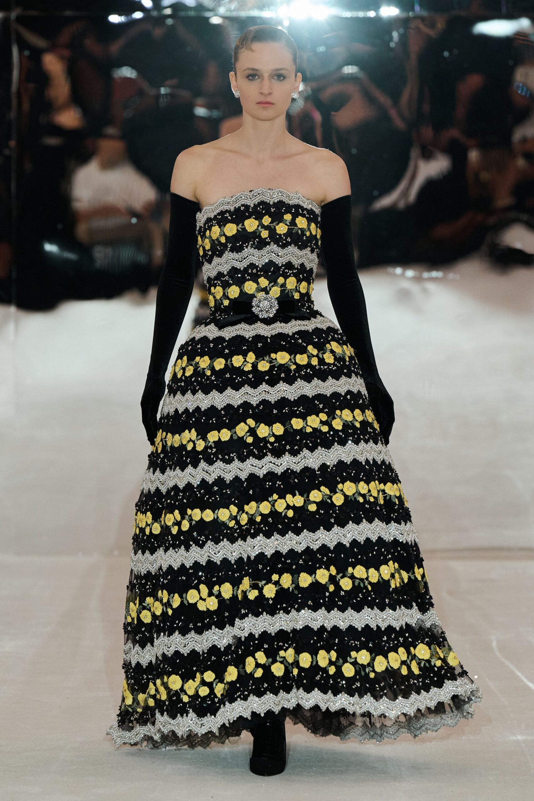 Strapless black ballgown banded with silver zigzag trim and yellow floral embroidery. Black velvet belt centered by a clustered crystal buckle. Long black velvet gloves disappearing into the dark skirt volume. Crystal flower studs. At the charity auction after midnight, still standing under the ballroom chandeliers.