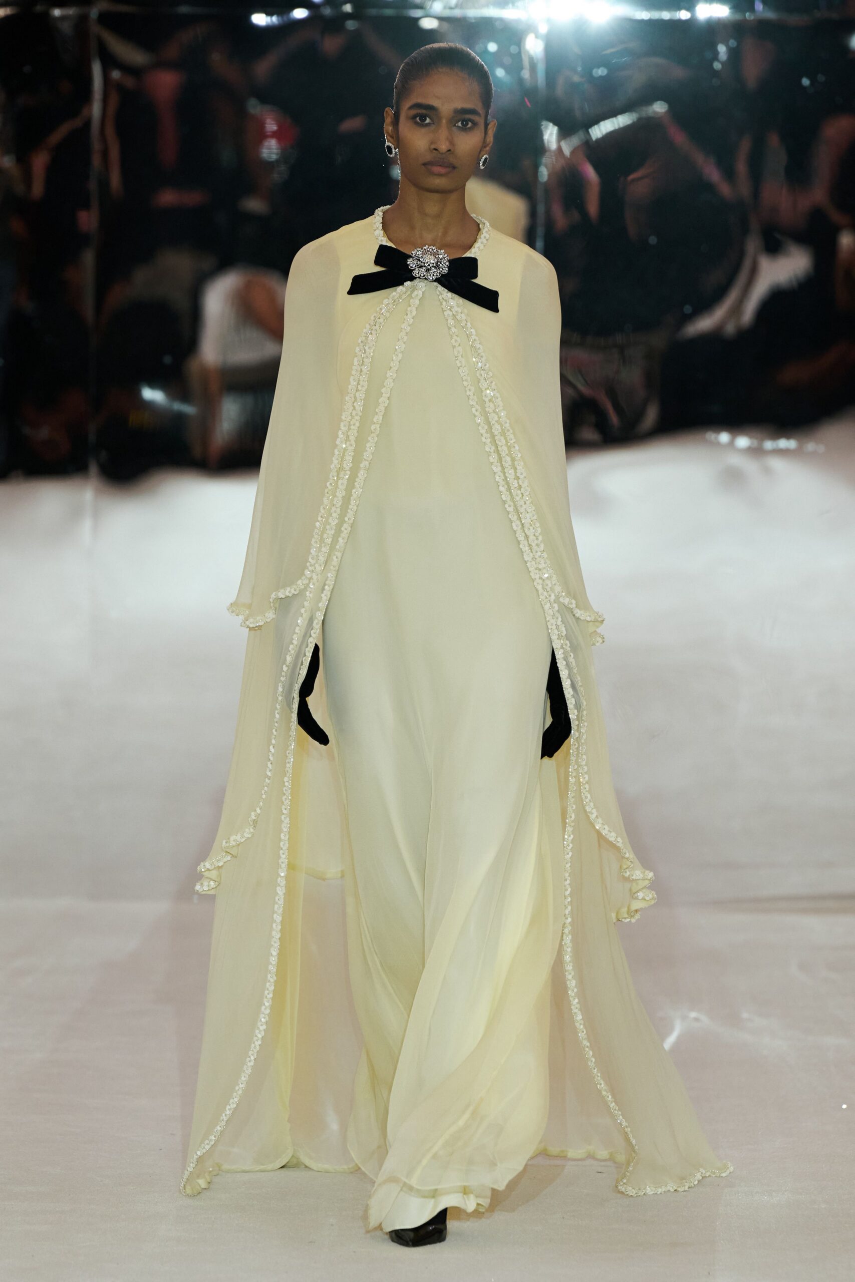 Crystal cluster brooch pinned over a flat black velvet bow. Buttercream chiffon cape gown, pearl-beaded edges falling in long open panels. Black opera gloves disappearing beneath the draped sides. Silver oval earrings with a dark center. For slipping down a hotel staircase after the orchestra has packed up.