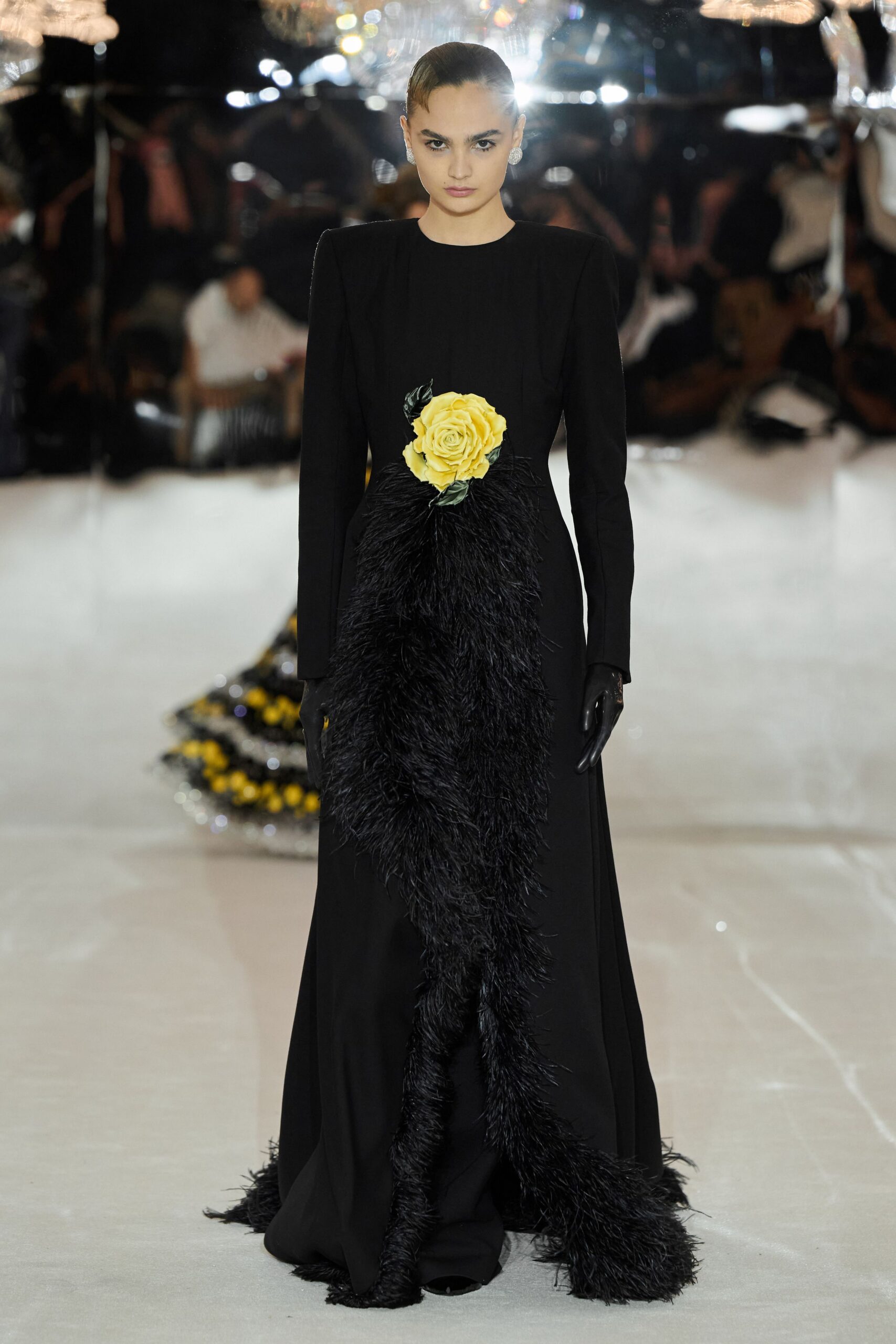 Round crystal clip earrings, bright as coins at each ear. Black floor-length column gown with hard shoulders and a yellow rose at the waist. Shaggy black feather drape falling from the rose to the hem. Glossy black opera gloves. Crossing a hotel ballroom after the funeral flowers were moved aside.