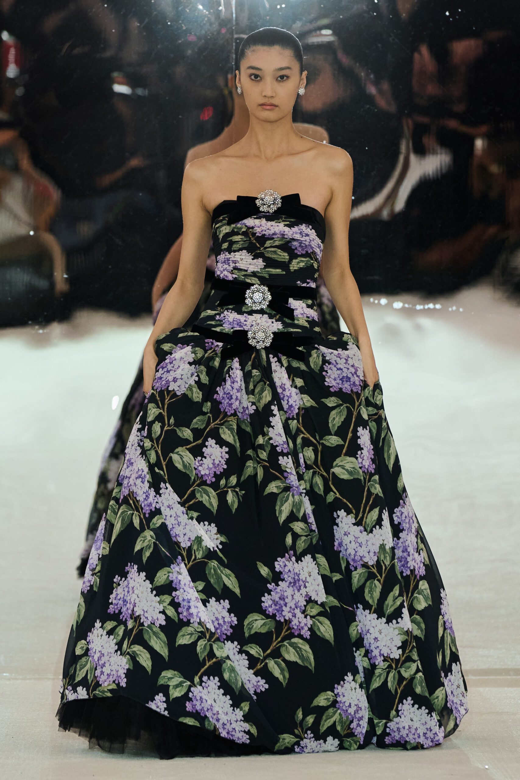 Strapless black ball gown printed with lilac sprays, velvet bows and crystal rosettes down the front. Small clustered crystal studs. Winter charity auction in a conservatory, orchids sweating against the glass.