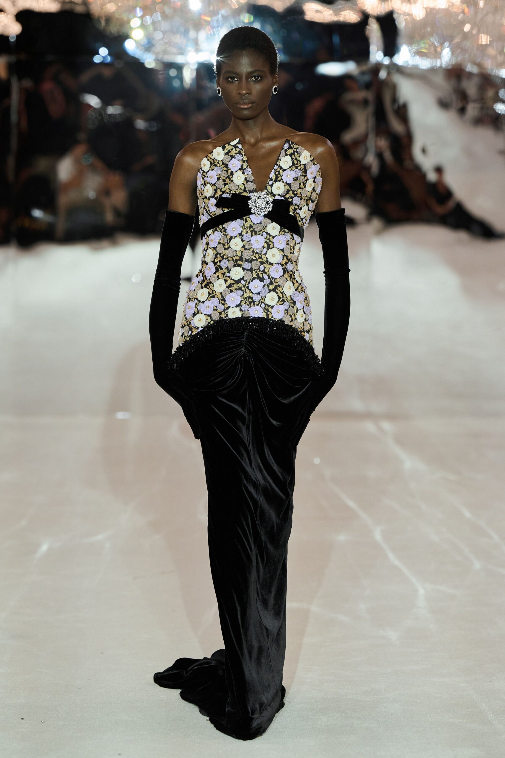 Floral brocade bodice with a black velvet band and crystal cluster. Black velvet column skirt, softly ruched at the hips. Black velvet opera gloves. Oval silver earrings. Under chandelier glare at a charity auction nobody can leave yet.