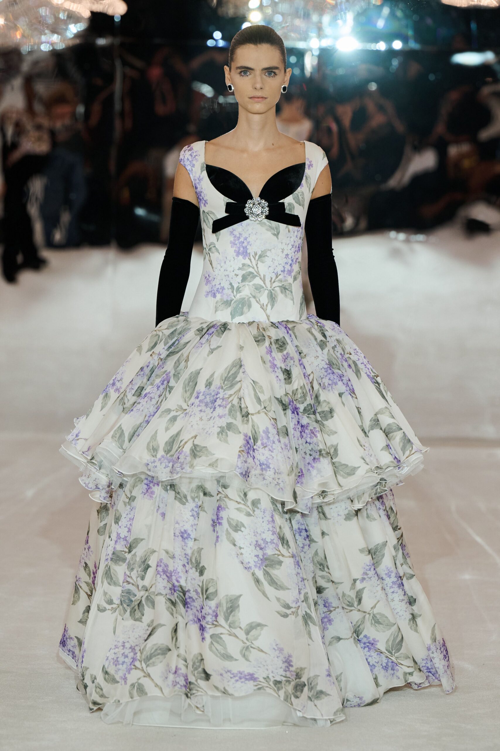 Black-and-silver hoop earrings. Opera gloves in black velvet. Ivory ball gown printed with lilac sprays, tiered organza flounces and a crystal brooch at the bow. Crossing a hotel ballroom before the string quartet starts.