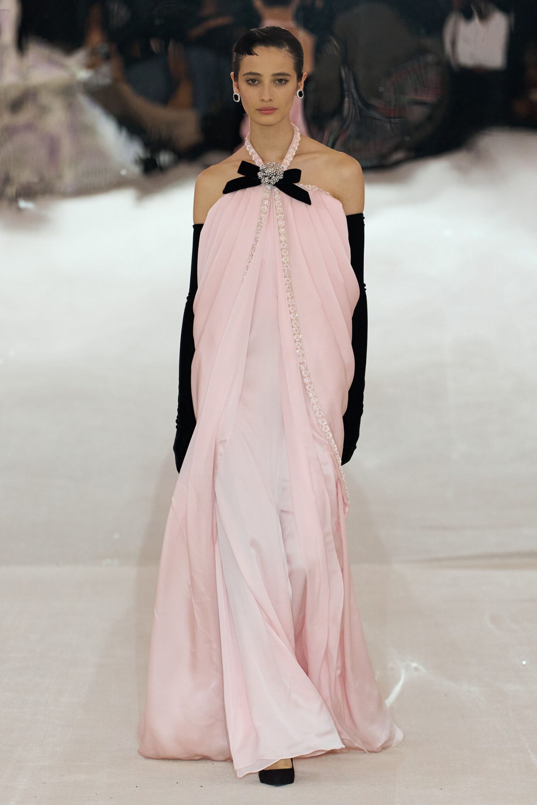 Blush chiffon gown draped from a jeweled rosette with a black velvet bow. Black opera gloves in matte stretch jersey. Oval crystal earrings with dark centers. Black square-toe pumps peeking from the hem. Crossing a hotel ballroom after midnight with confetti still underfoot.
