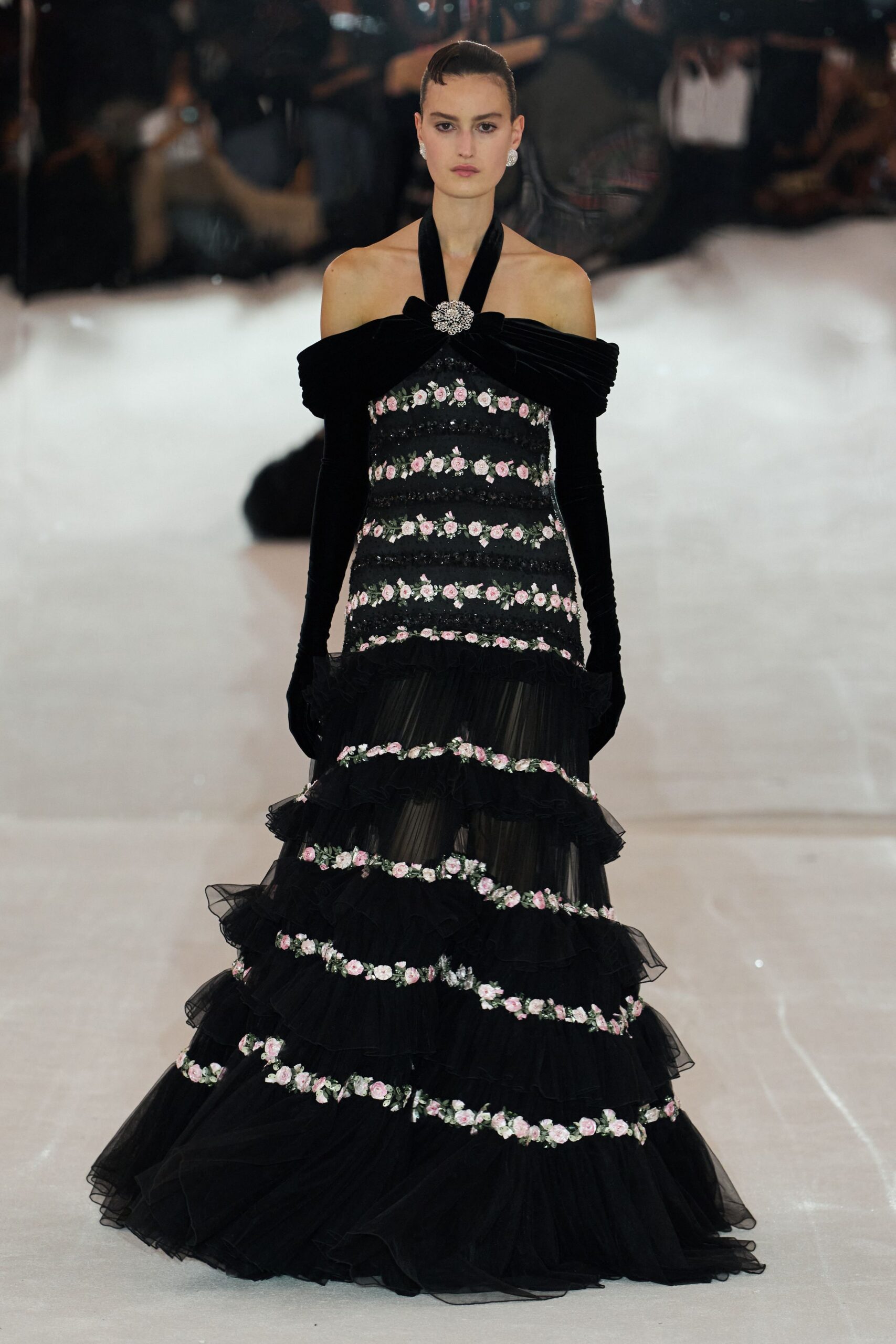 Round crystal cluster earrings. Black evening gown with velvet off-shoulder twist, rhinestone brooch, and pink rose garlands over tiered tulle. Opera-length black velvet gloves disappearing into the sleeves. At a charity ball in an old conservatory after the rain stopped.