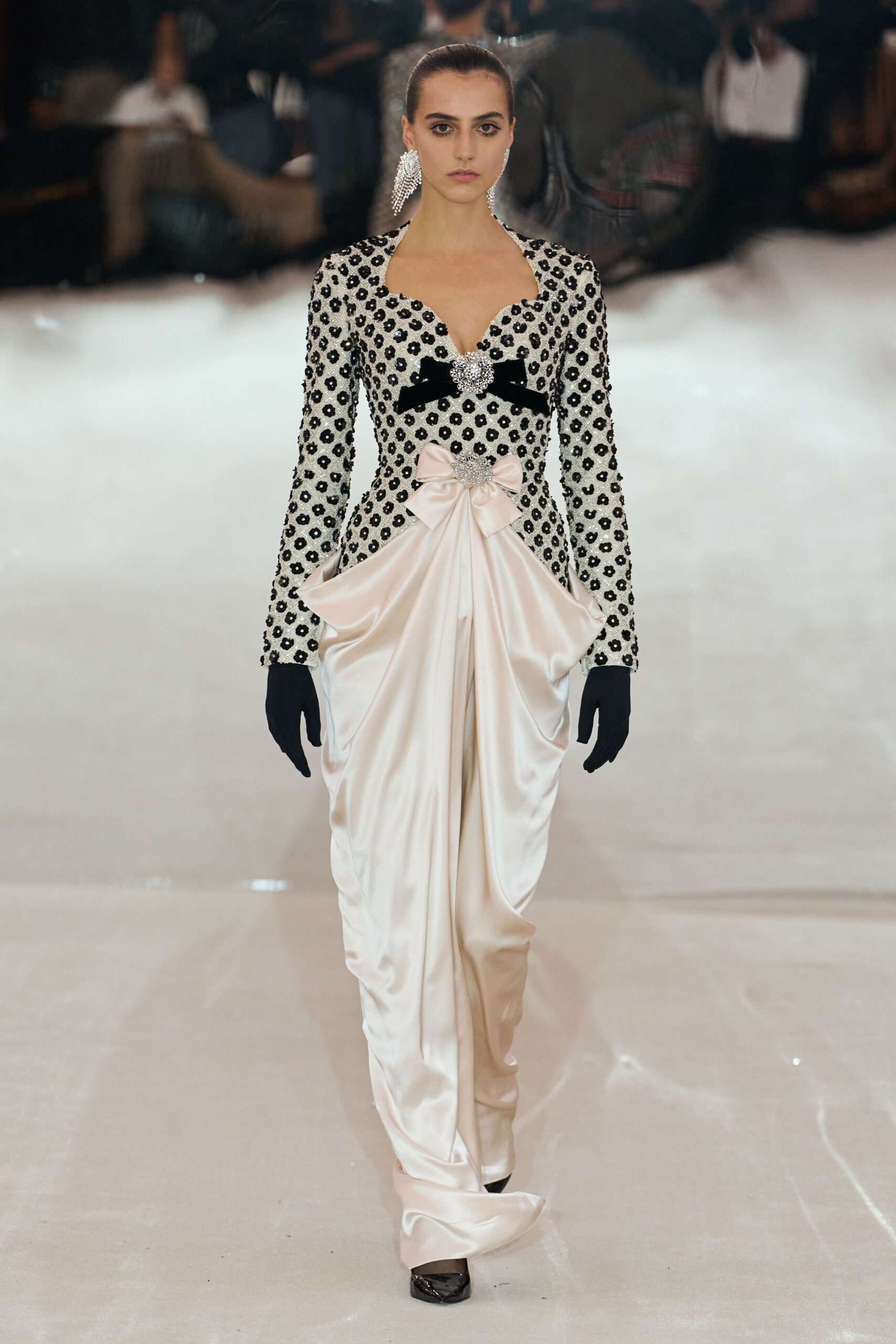 Ivory satin gown with a black flower-beaded bodice and pooled draped skirt. Black velvet bow pinned with a crystal cluster at the bust. Blush satin bow cinching the waist, slightly off center. Black opera gloves. Chandelier crystal earrings. At the charity ball after the orchestra has packed up its cases.
