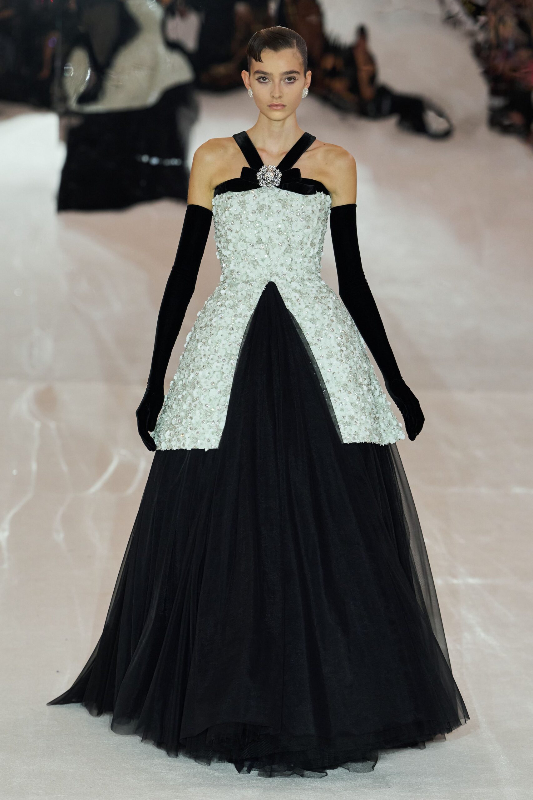 Ivory beaded bodice over a black tulle skirt, front split in a dark wedge. Black velvet cross straps pinned with a crystal rosette. Long black gloves in matte stretch velvet. Round crystal cluster studs. The final toast in a ballroom after the quartet has packed up.