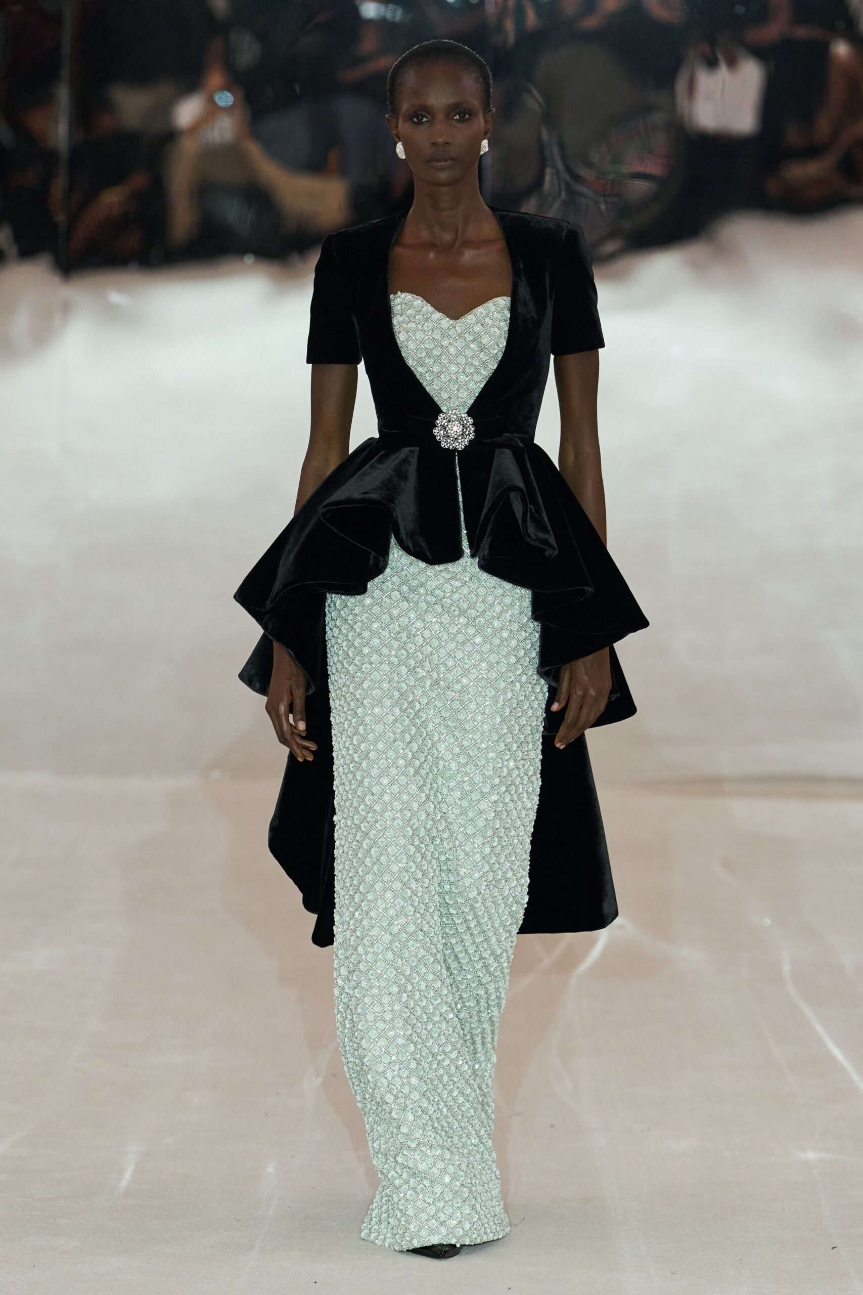 Round crystal cluster earrings. Ice-blue beaded column gown, diamond lattice catching light across the body. Black velvet short-sleeve overskirt, cinched with a rhinestone brooch at center. For arriving at a hotel ballroom after the orchestra has already started.