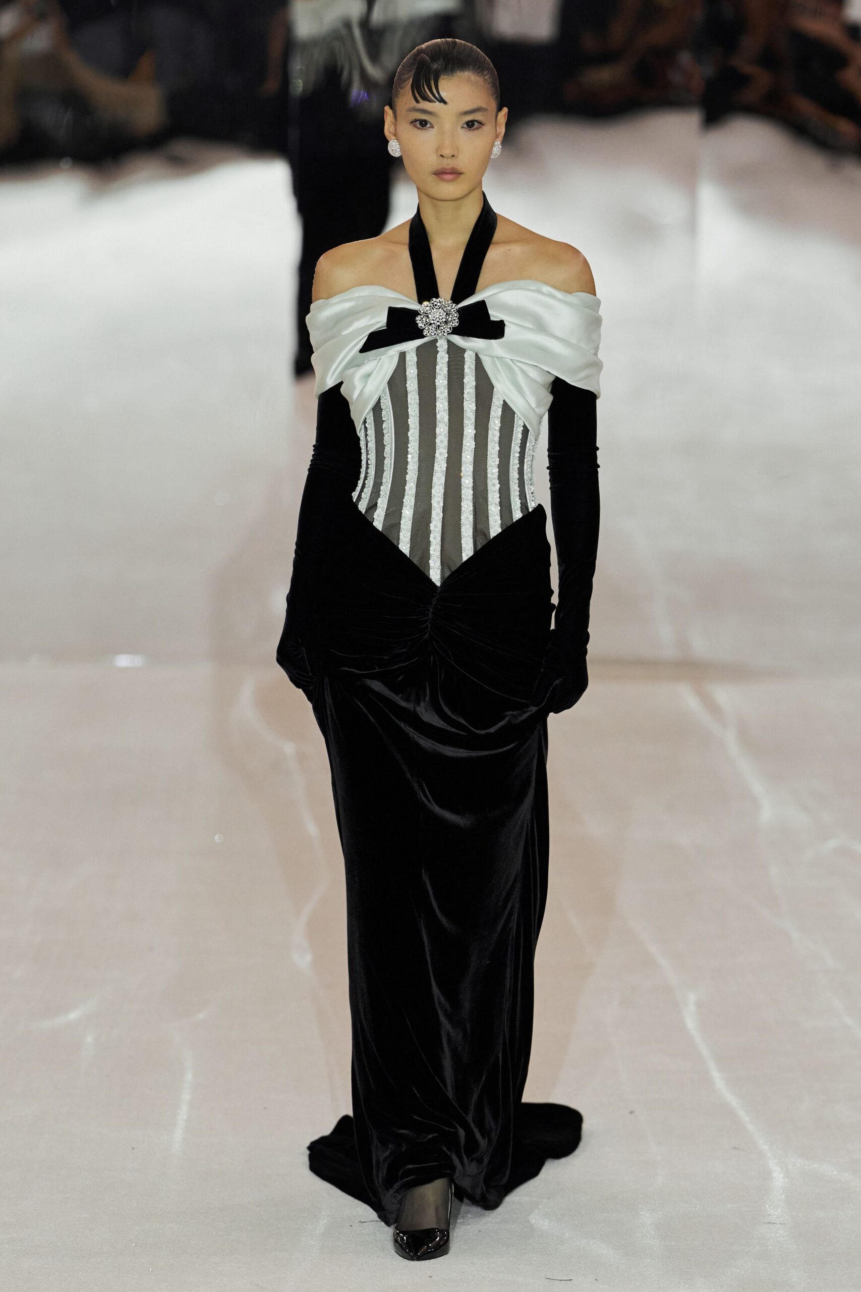 Ivory satin off the shoulder, pinned with a crystal brooch and black bow. Sheer dove corset striped with sequined channels. Black velvet opera gloves disappearing into the bodice. Black velvet column skirt, ruched hard at the hips. Patent pointed pumps. For crossing a hotel ballroom after the quartet has packed up.