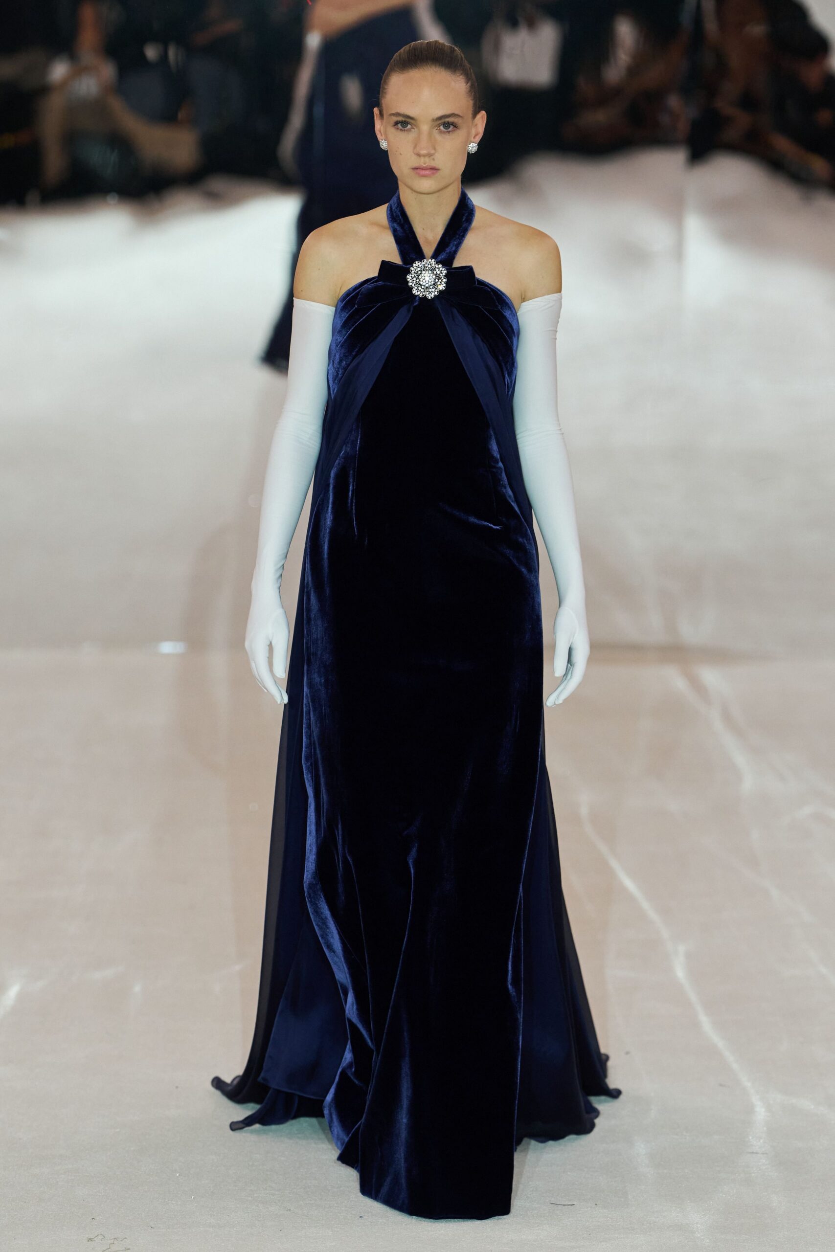 Midnight velvet halter column gown, rhinestone brooch pinching the draped neckline. White stretch opera gloves, stark against the bare shoulders. Cluster crystal studs. Crossing a hotel ballroom after the orchestra has packed away.