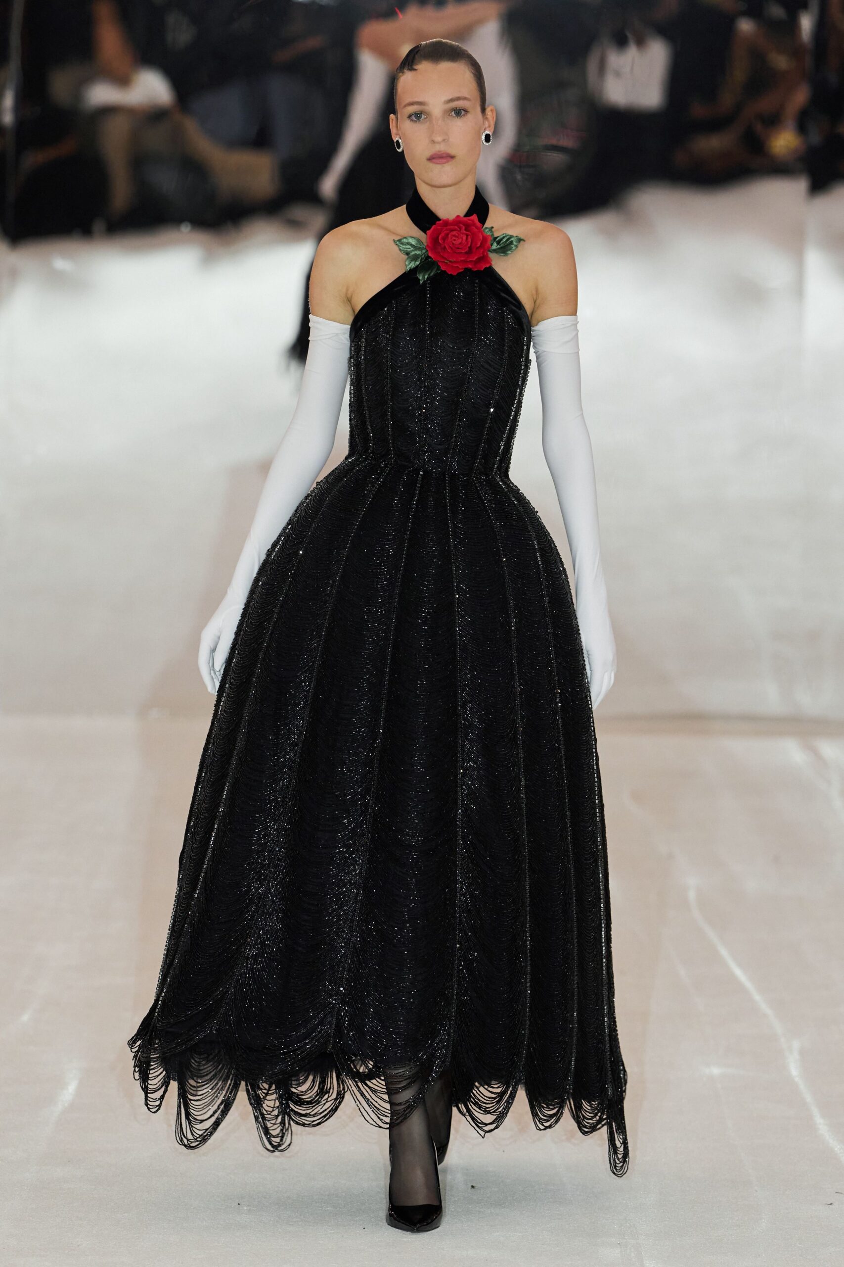 Black halter ball gown crusted in beading, skirt hung with looped fringe swags. Red rose corsage with two green leaves fixed at the throat. White opera gloves slipping just below the shoulders. Sheer black tights. Black pointed pumps. At the winter charity gala after someone opened the terrace doors.