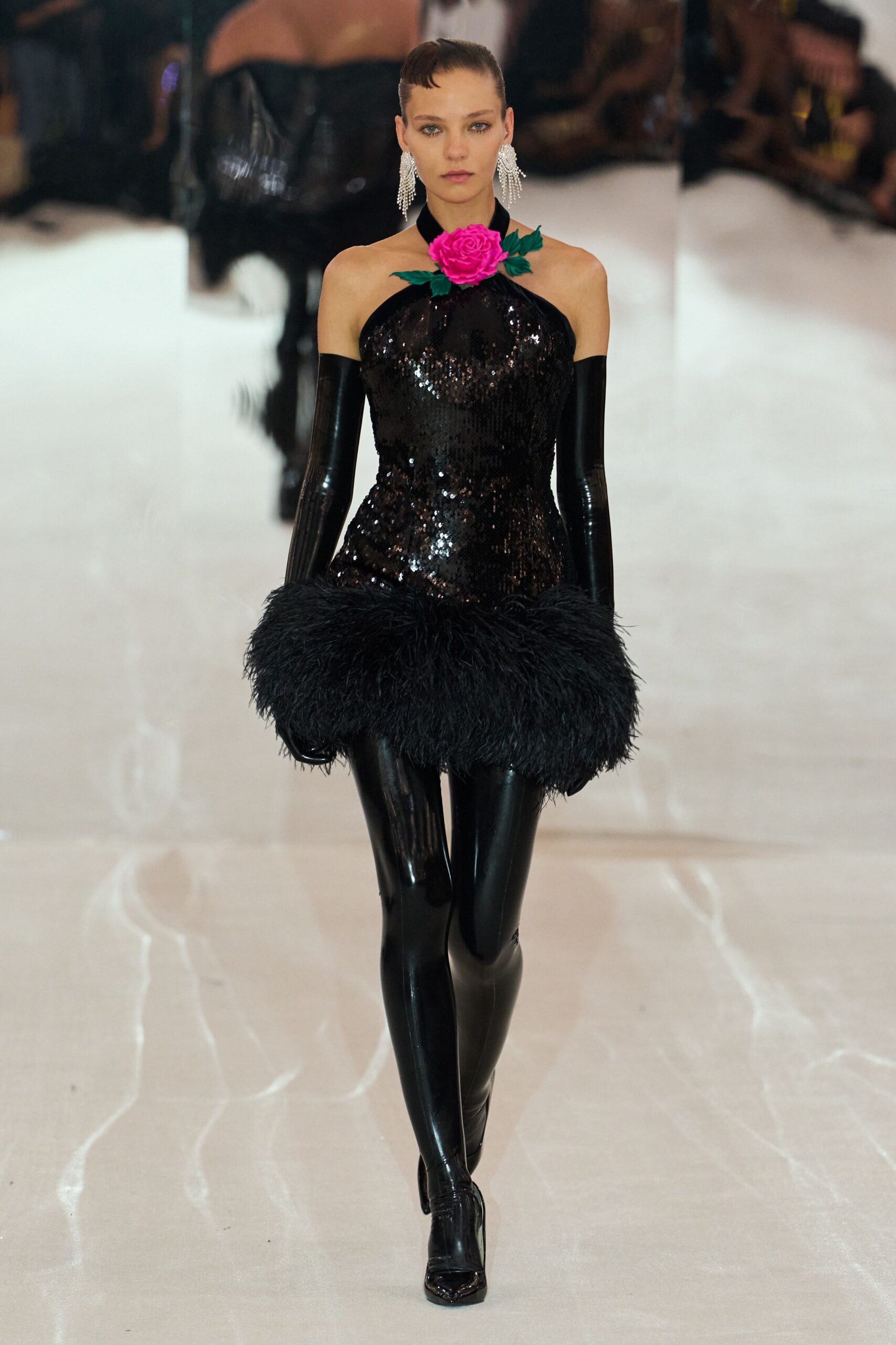 Crystal fringe earrings brushing the jaw. Black sequined halter mini with a hot-pink rose pinned at the throat. Glossy black gloves ending in shaggy feather cuffs. Second-skin black latex leggings. Square-toe patent boots peeking under the shine. Hotel ballroom at midnight, somebody has knocked over the centerpieces.