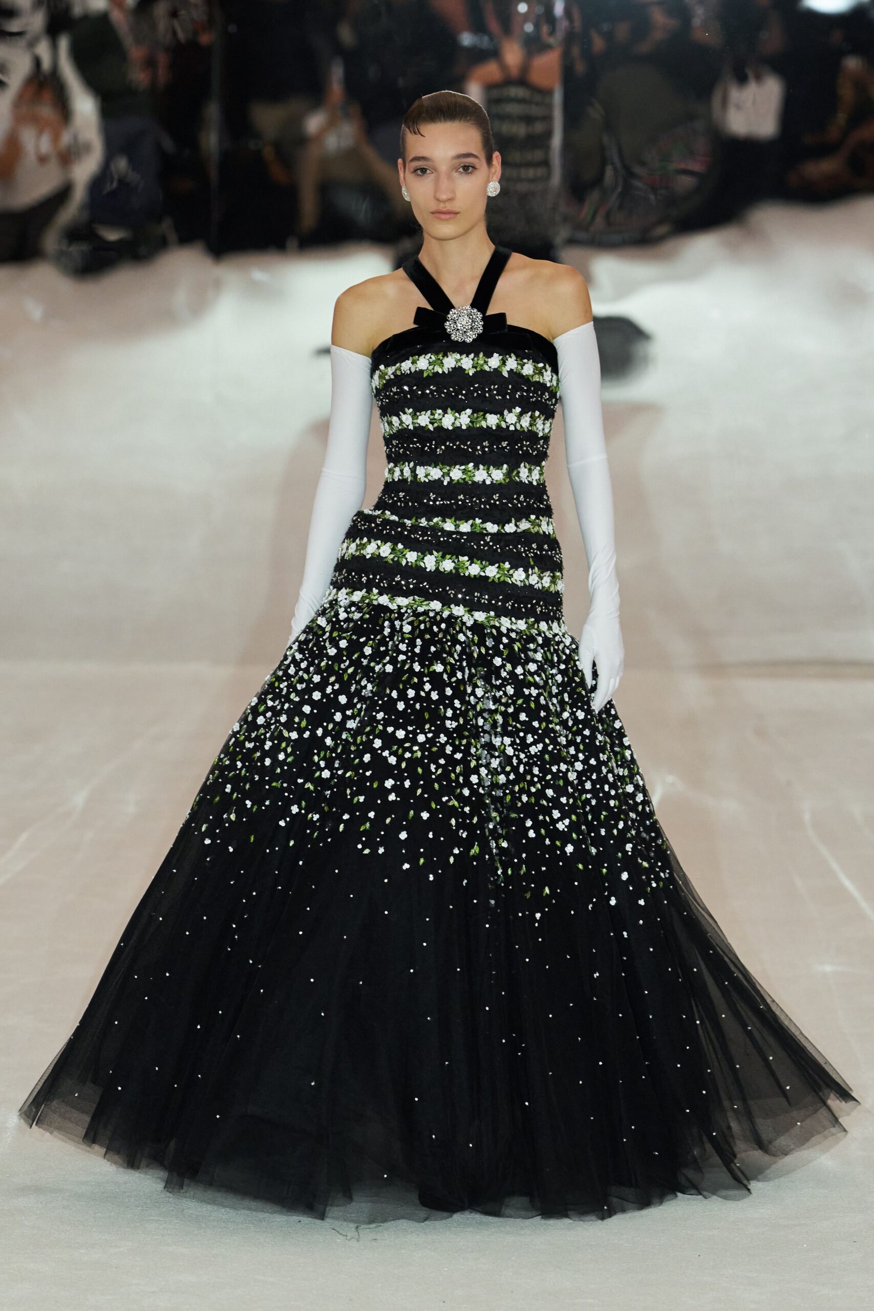 Round crystal cluster studs. White stretch gloves worn above the elbow. Black tulle ball gown banded with white floral embroidery, rhinestone brooch at the halter. Crossing a hotel lobby still full of wedding flowers after midnight.
