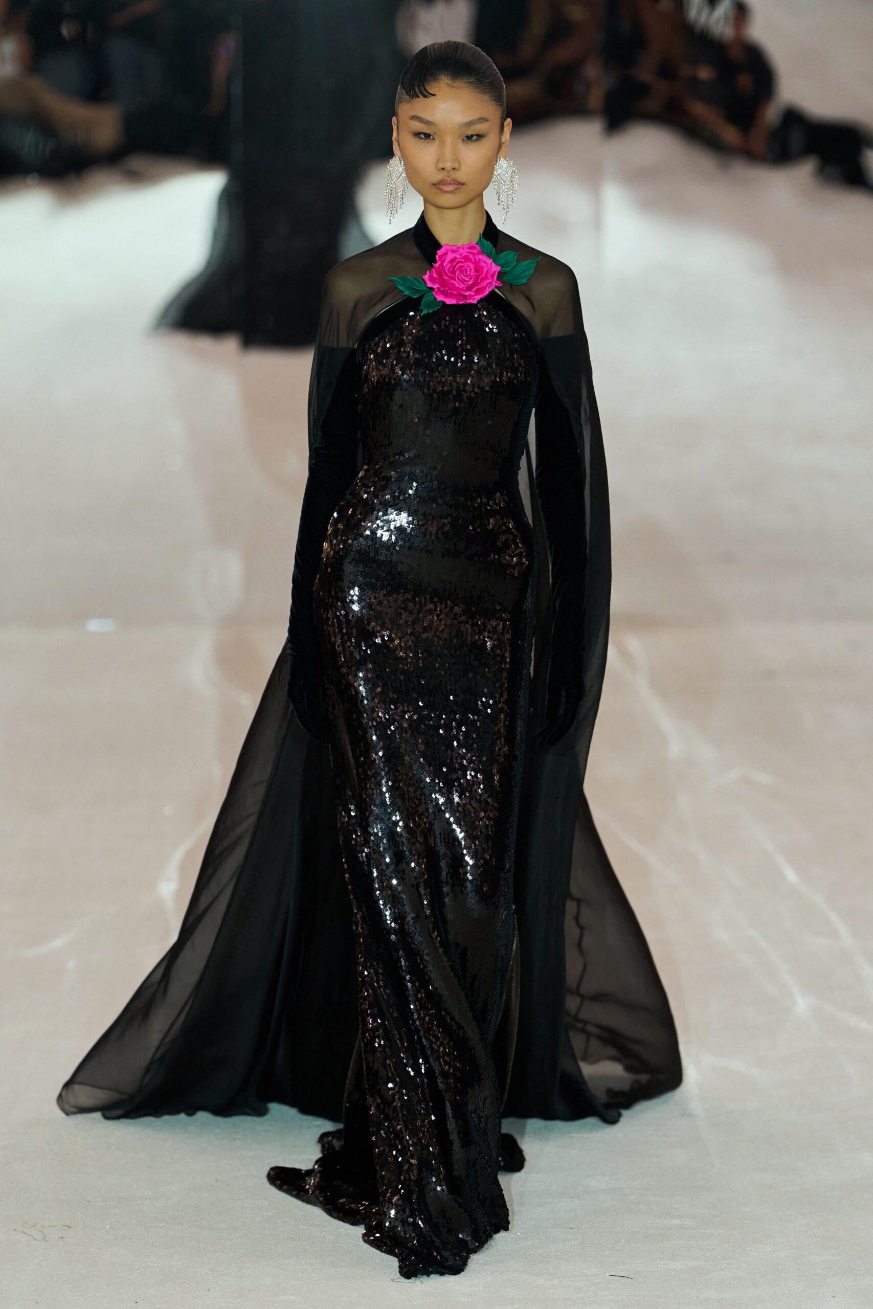 Crystal fringe earrings brushing the jaw in dense white strands. Black sequined column gown with a hot-pink rose pinned at the throat. Sheer black cape streaming from the shoulders behind velvet sleeves. Long black gloves, second-skin shine. Crossing a hotel ballroom after midnight with petals crushed underfoot.
