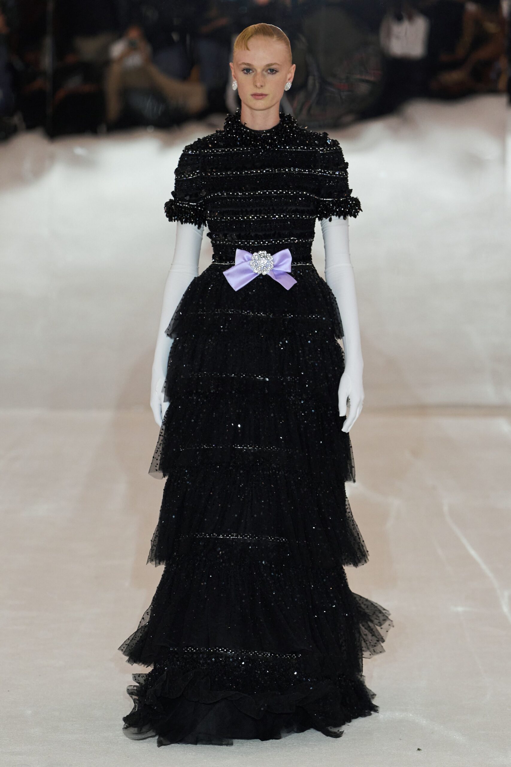 Black tulle evening gown, beaded cap sleeves and glittering tiered skirt. Lilac satin bow belt centered with a clustered crystal brooch. Opera gloves in stark white stretch satin. Round crystal clip earrings. At the hotel ballroom after midnight, when the cake is already cut.