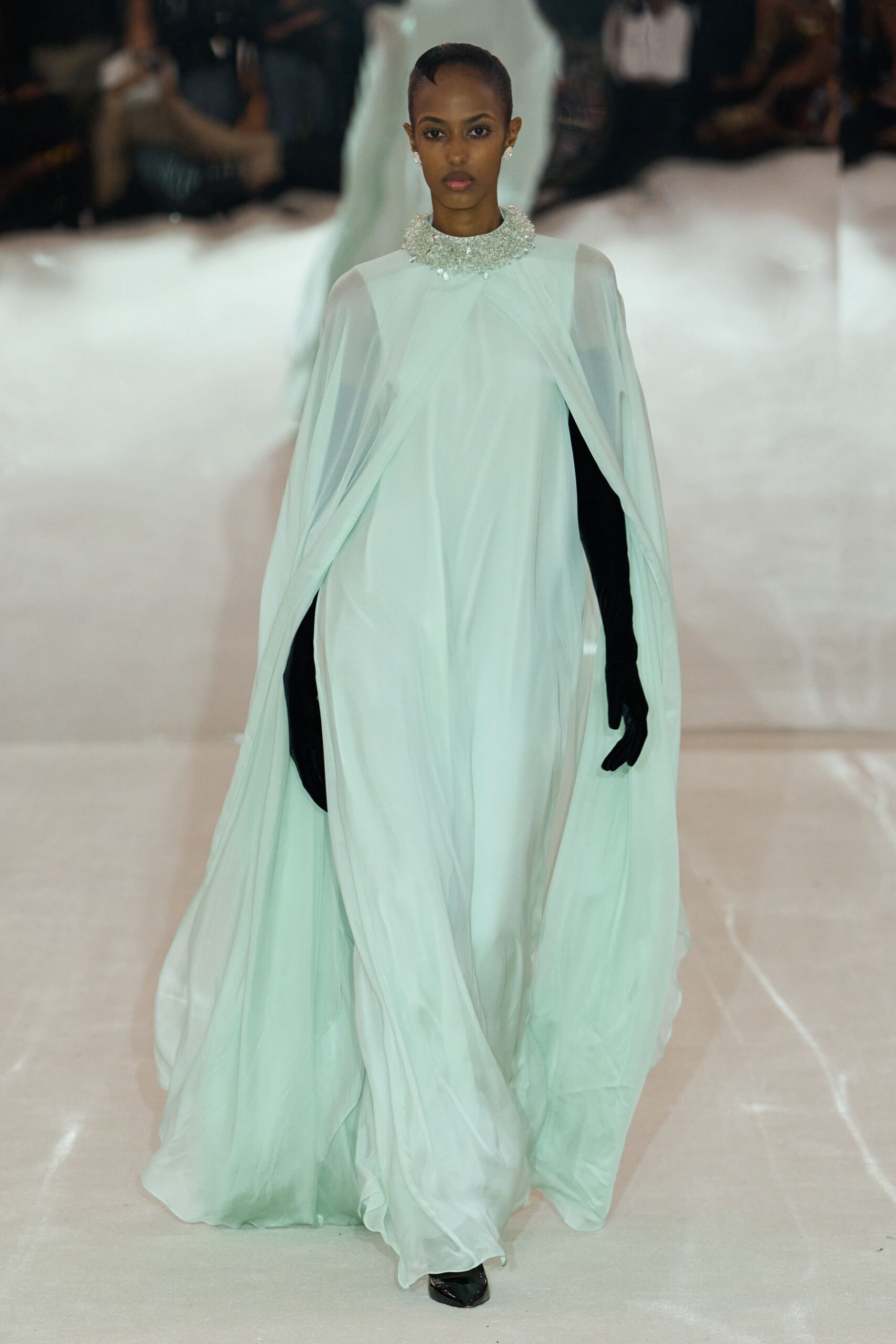 Pale mint chiffon gown, cape sleeves falling in long liquid folds. High collar crusted with clustered clear beads. Opera-length black gloves cutting a stark line under the drape. Black pointed shoes barely breaking the hem. Crossing a hotel ballroom at dawn while the ice sculptures melt.