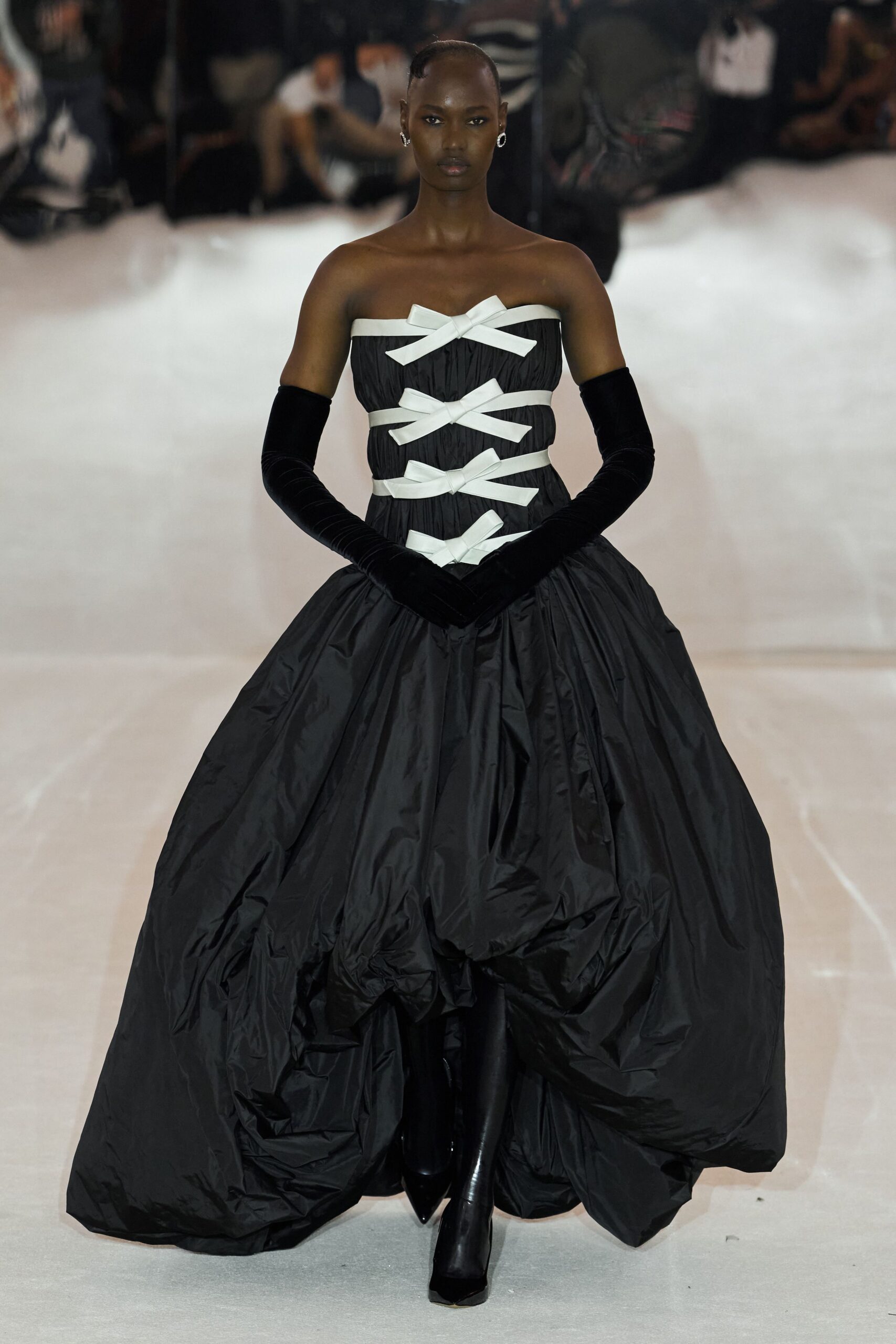 Strapless black taffeta ballgown, corseted with four white grosgrain bows. Black velvet opera gloves reaching above the elbow. Black pointed boots disappearing under the ballooned hem. Small silver hoop earrings. At the charity auction after someone opens the sealed music room.