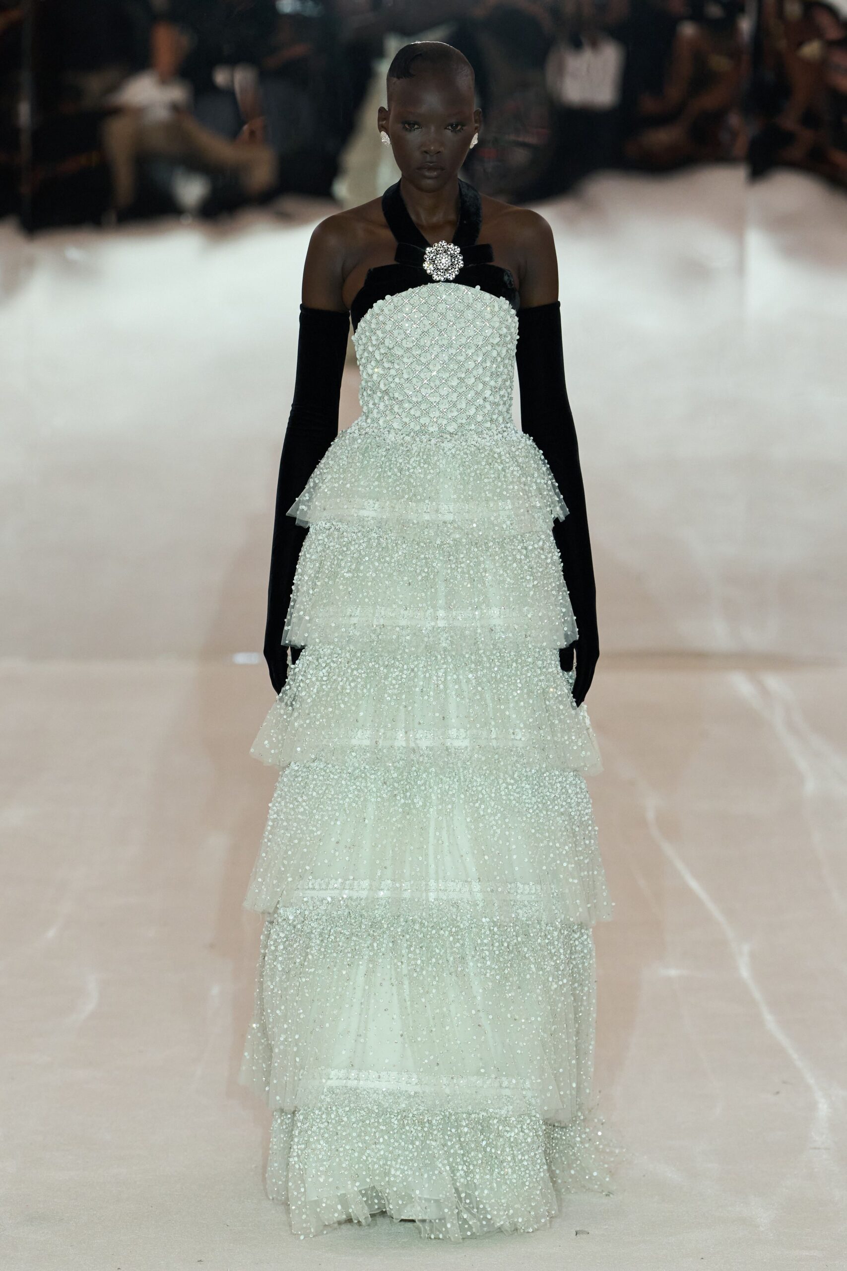 Small crystal cluster studs. Ivory tulle column densely beaded, stacked in wide glittering tiers. Black velvet halter band crossed with a rhinestone brooch. Long black velvet gloves disappearing beneath the bodice. At the hotel ballroom after midnight, when the ice sculpture starts to melt.